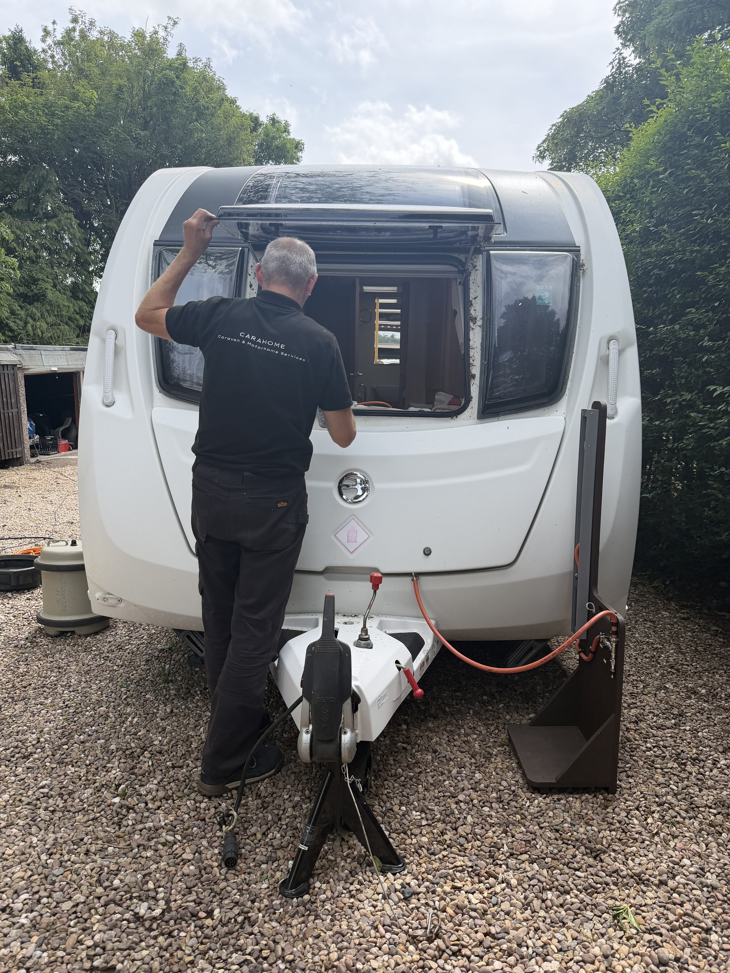 A person working on the front of a travel trailer with a black shirt that reads 'Carahome' and 'caravan & motorhome services,' outdoors on a gravel surface near trees, with equipment and tools nearby.