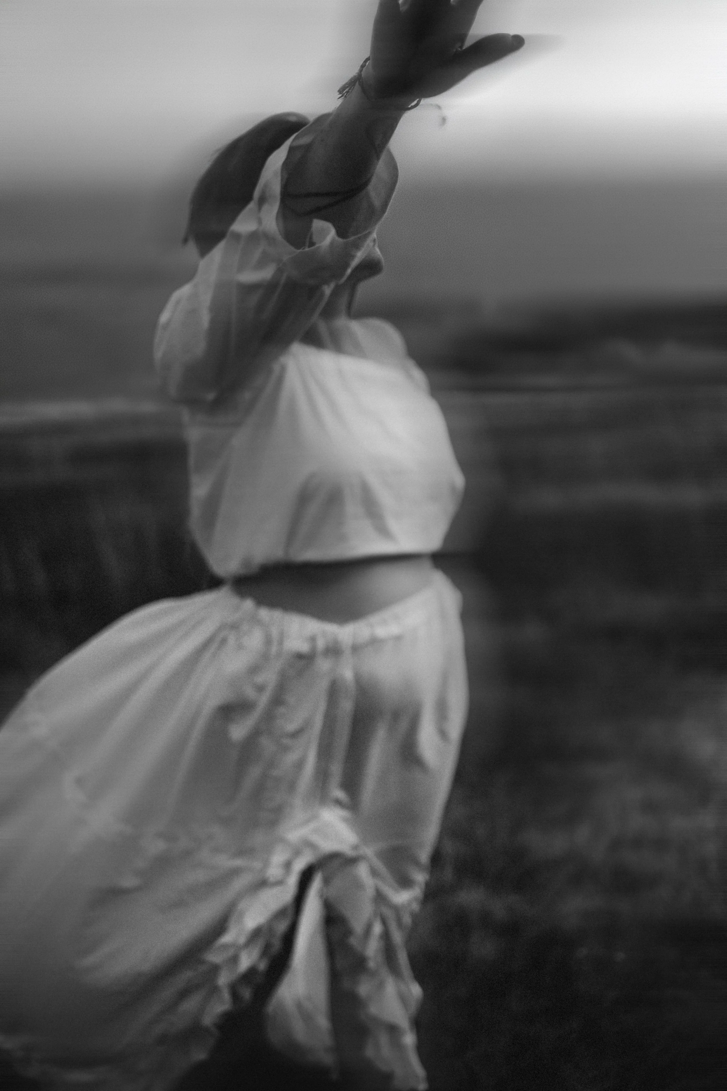 A girl near the beach, reaching upward with her right hand, wearing a white crop top and light-colored pants, with blurred background