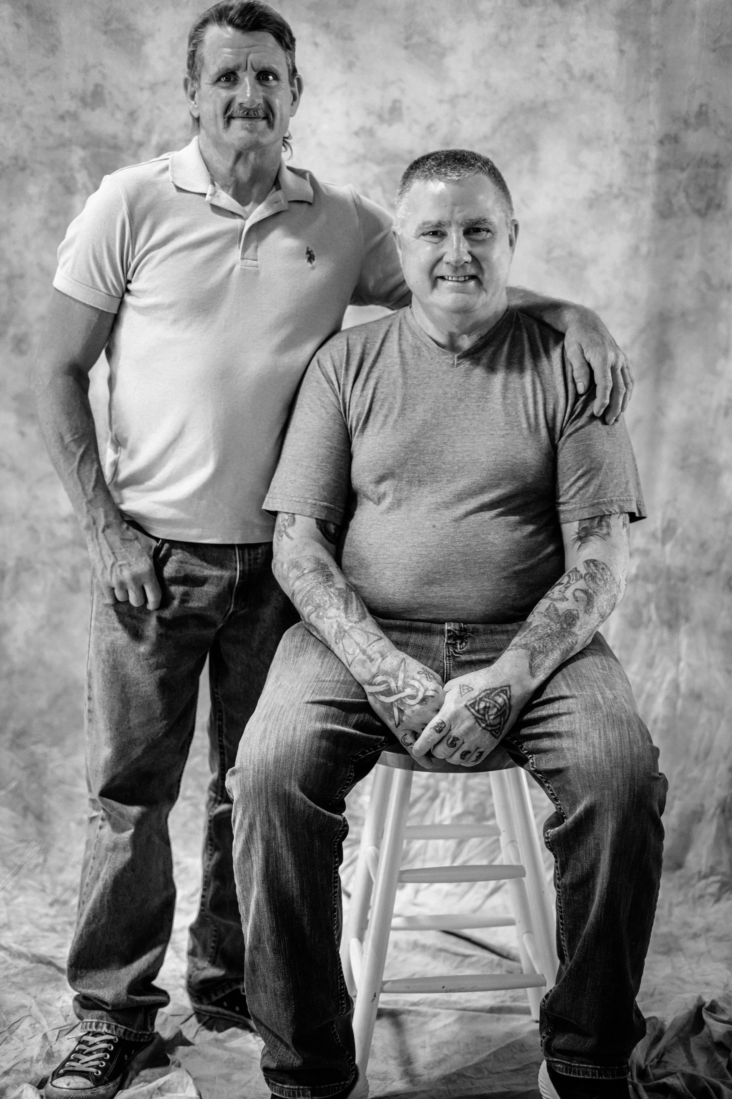 Two men in casual clothing, one standing with his arm around the other's shoulder, both smiling at the camera, in a studio with a plain background.