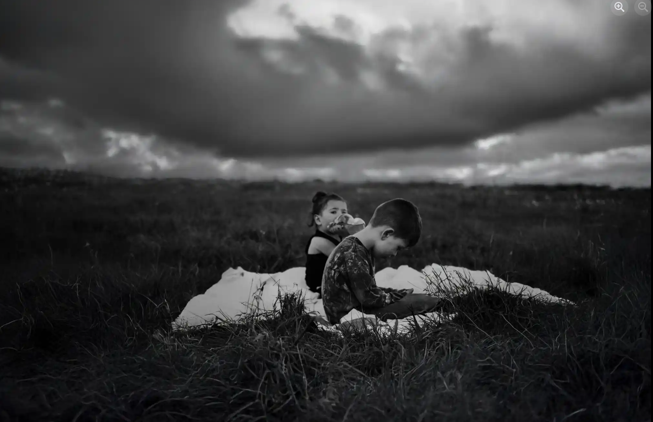 Two children sitting on a blanket in a vast field under a dark, cloudy sky, with one child eating and the other looking at a device, in black and white.