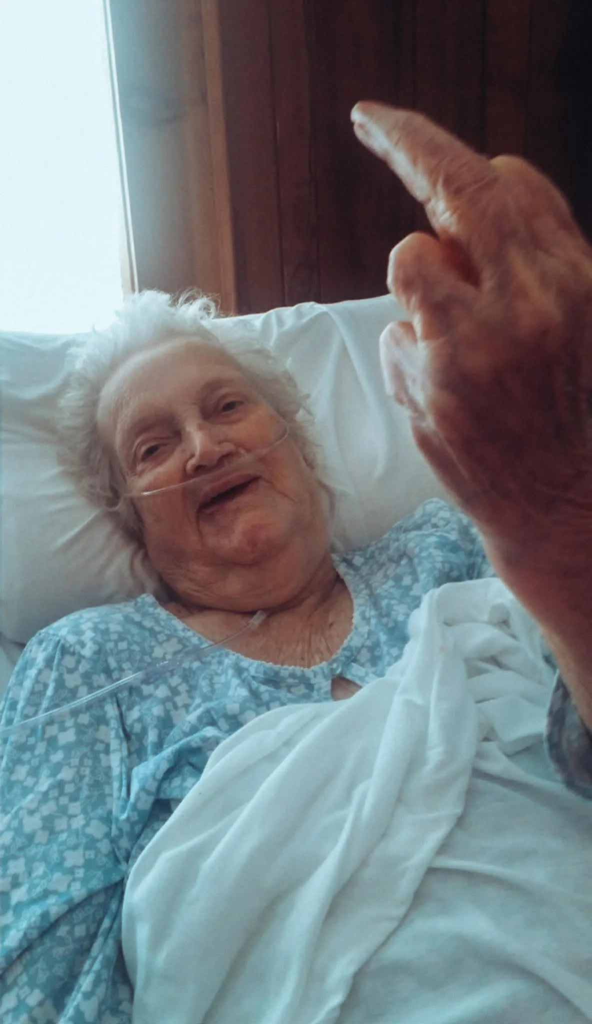 An elderly woman lying in bed with a nasal oxygen tube, smiling and looking at a dog with a paw raised, in a room with wood-paneled walls.