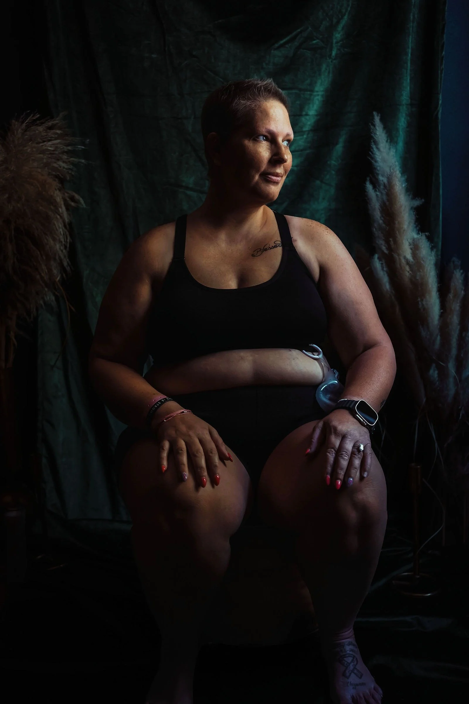 A woman with short hair, wearing a black sports bra, sitting on a chair in a dark setting. She has a tattoo on her chest, a smartwatch on her left wrist, and rings on her fingers. The background includes dark drapery and some pampas grass.
