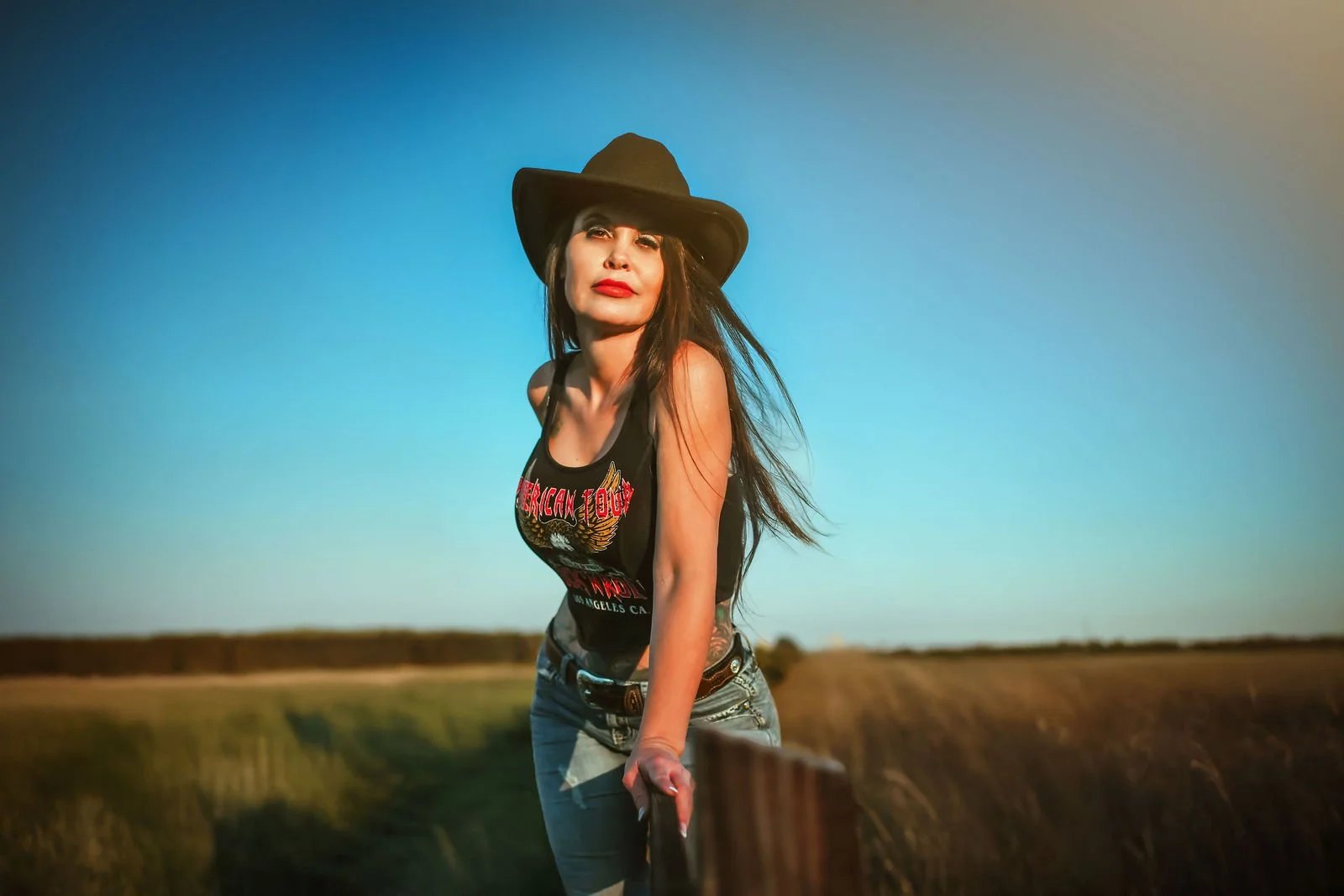 A woman with long brown hair wearing a black cowboy hat, red lipstick, a black tank top with a graphic print, and blue jeans, standing outdoors in a rural setting with fields and a clear sky.