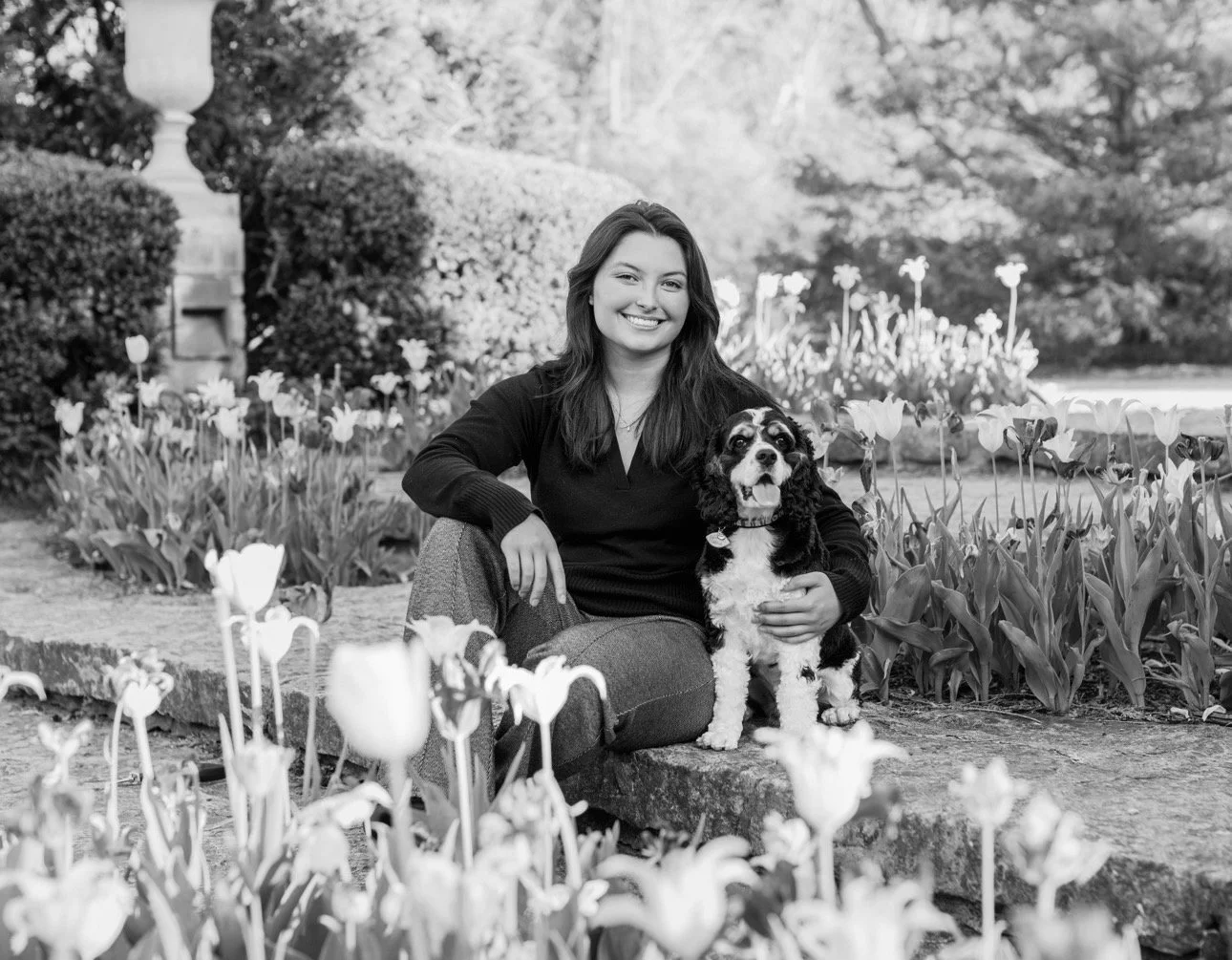 A woman sitting on a stone ledge in a garden with flowers, holding a dog.