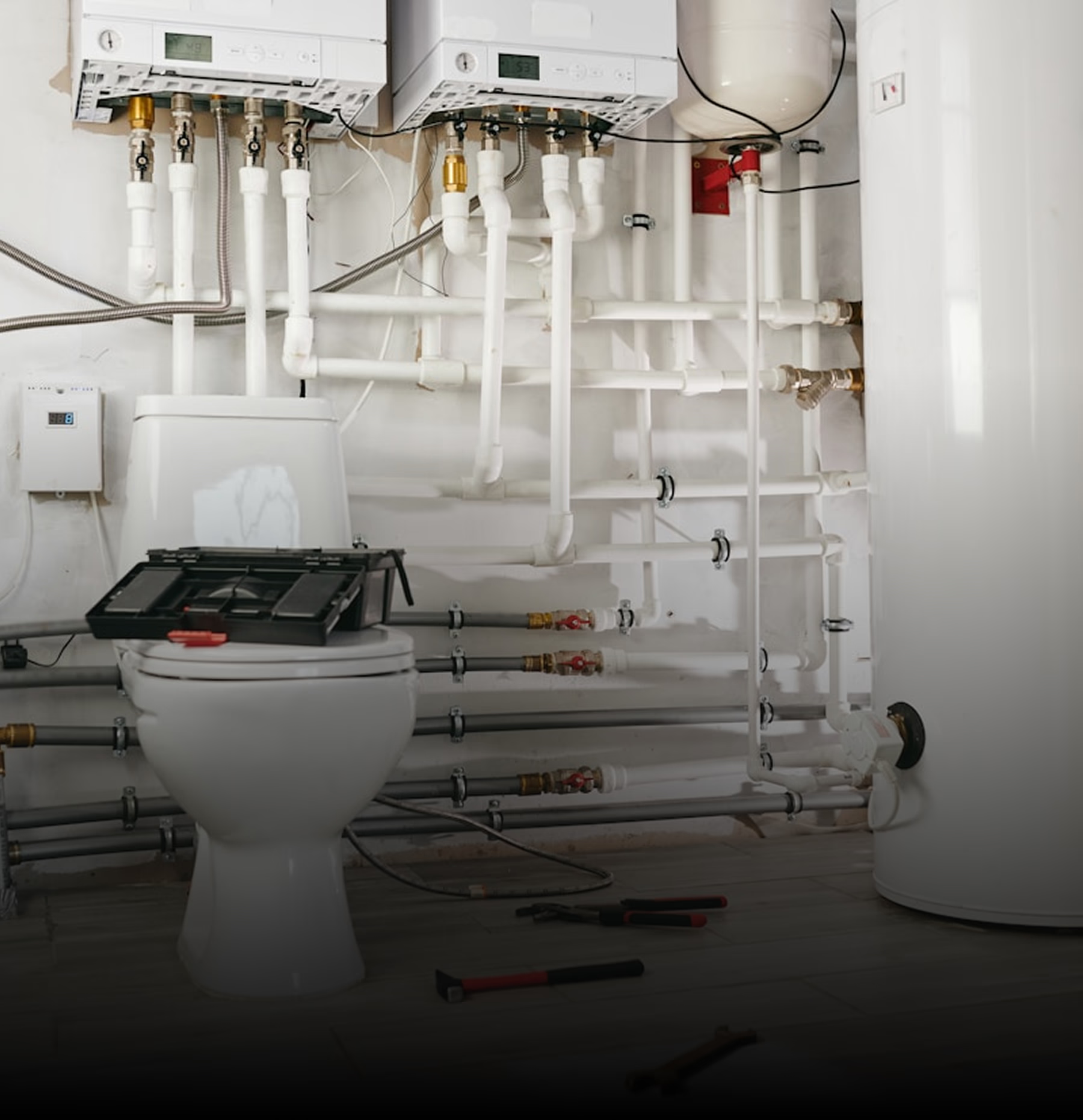 Close-up view of plumbing pipes and two water heaters installed on a wall, with tools on the floor.