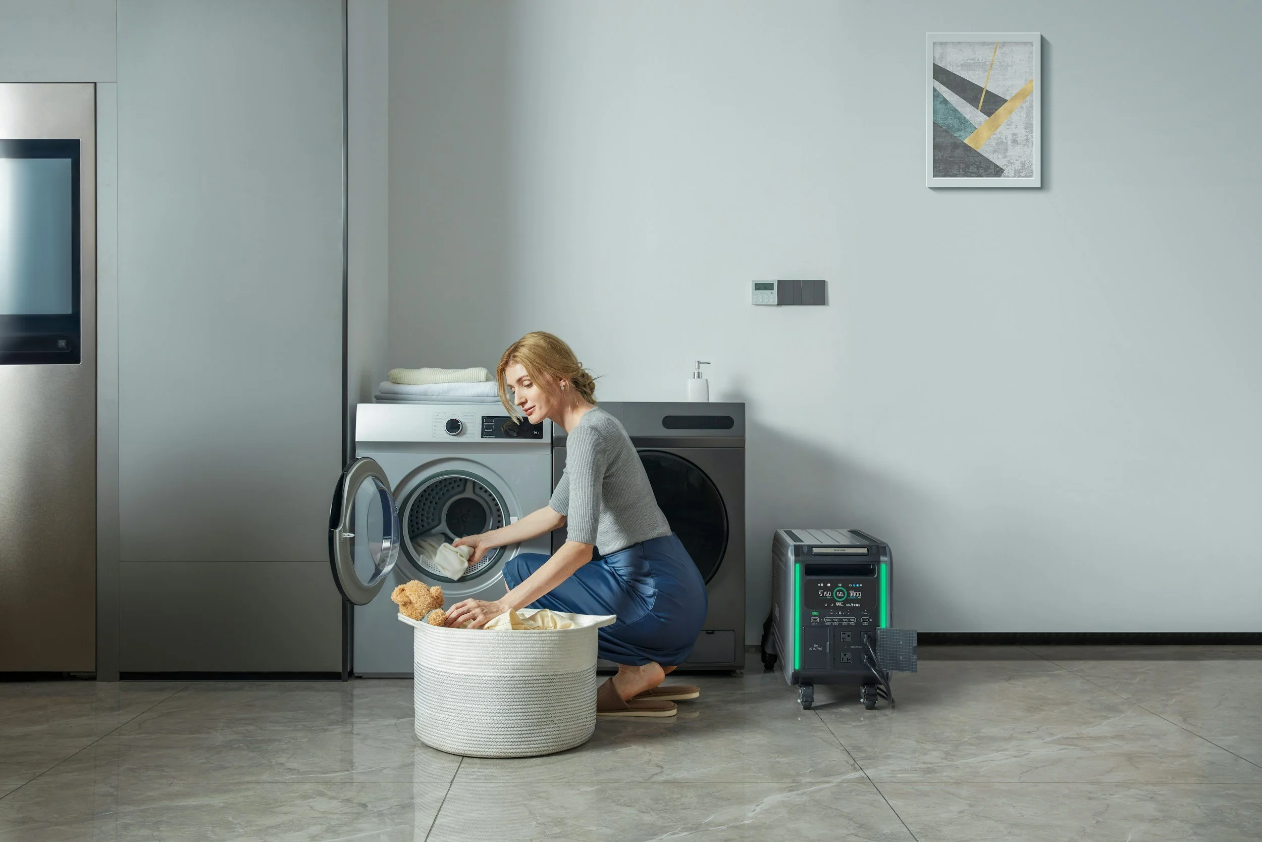 A woman crouching in a laundry room, loading clothes into a front-loading washing machine. She has short blonde hair and is wearing a gray top and blue pants. There is a laundry basket with a teddy bear and clothes in front of her, and a white container with a soap dispenser on top. Behind her are stacked towels, a wall art with abstract shapes, and a wall-mounted thermostat. To the right, there is a portable air purifier with green accents.
