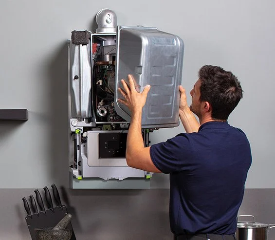 A technician working on a wall-mounted boiler or water heater, opening its metal cover for repair or inspection.
