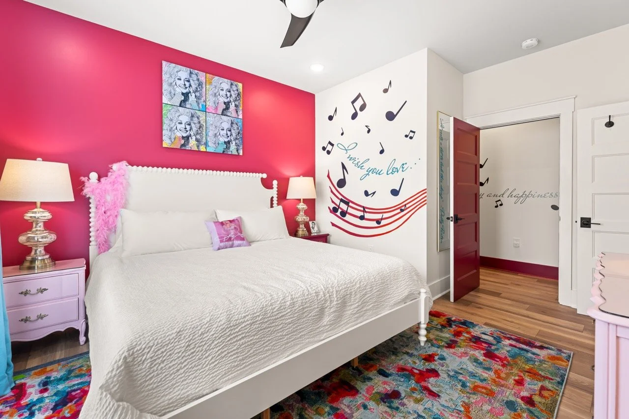 A bedroom with a white bed and a pink wall behind it. The wall has a colorful painting of a smiling woman and musical notes with the text 'wish you love.' There are pink nightstands with lamps on each side of the bed, a rainbow-colored rug, and a woo