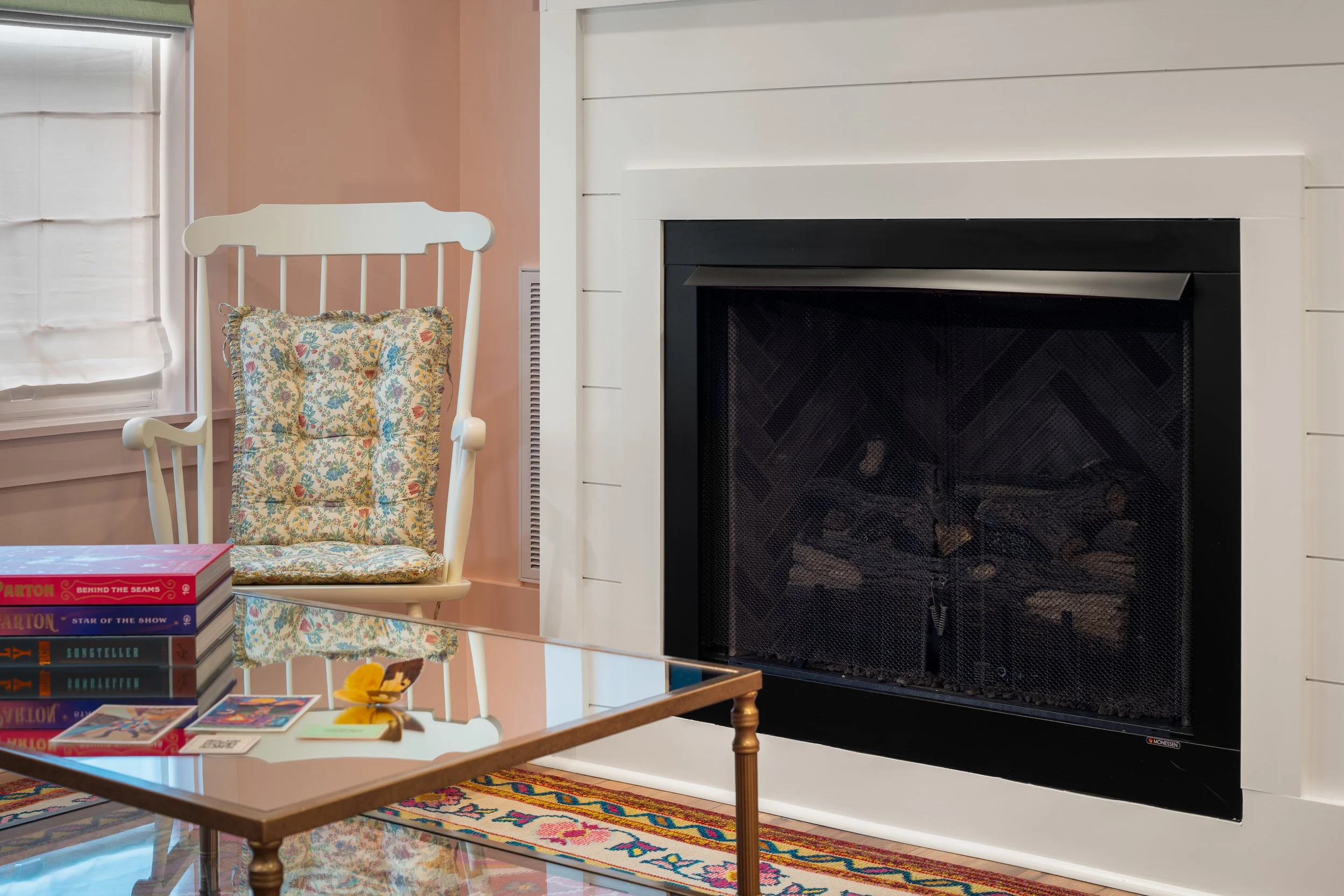A cozy living room corner with a white rocking chair decorated with a floral cushion, beside a fireplace with a white mantel, and a window with a white curtain. A coffee table with stacked board games and a small decorative object is in the foregroun