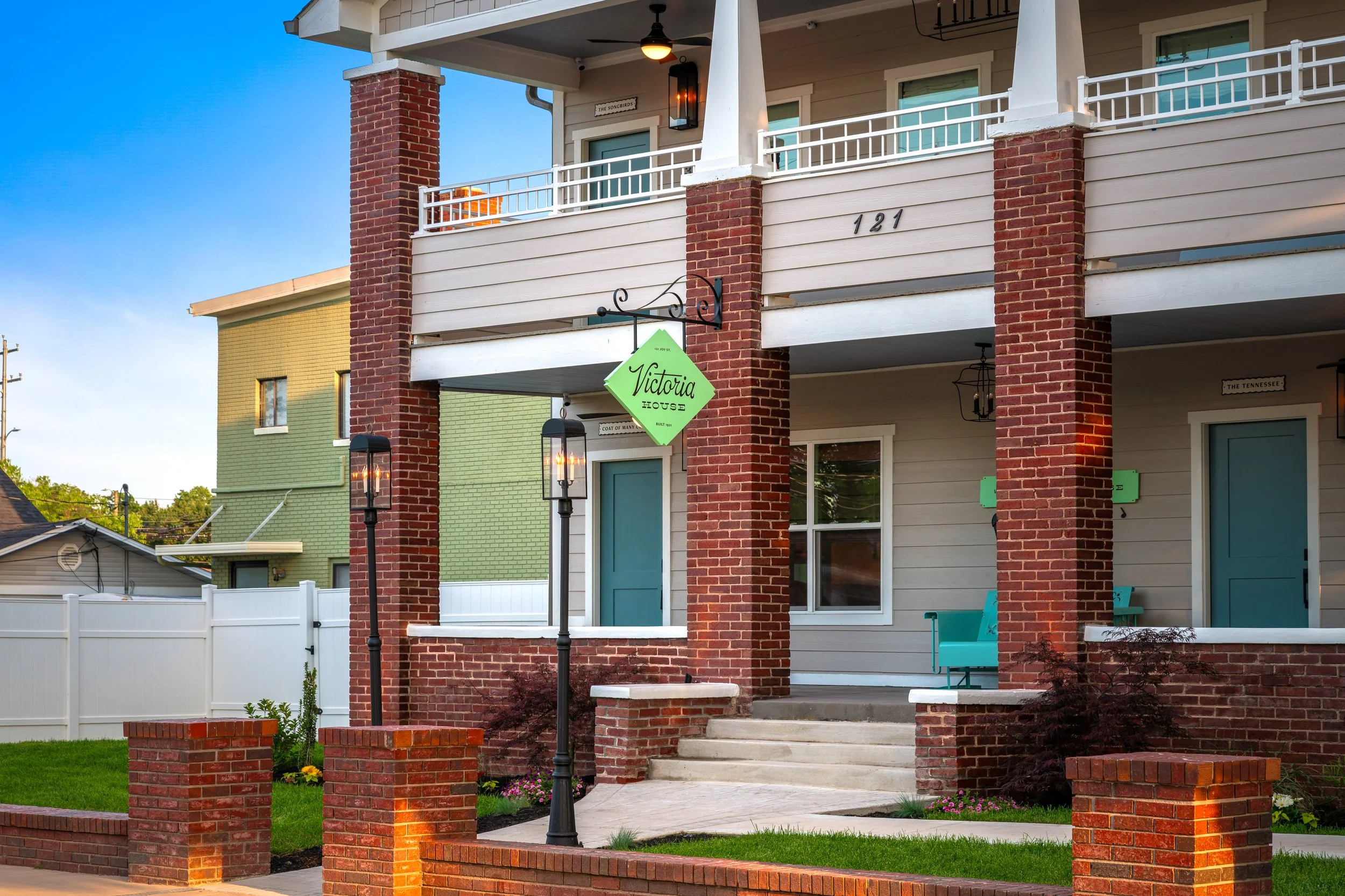 Front porch of a house with brick pillars, a green sign that reads 'Victoria House,' stairs, blue chairs, and a white fence in the background.