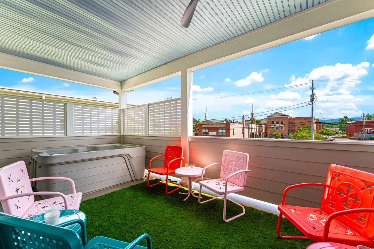 Outdoor balcony with colorful chairs, a hot tub, artificial grass, and a city view under a blue sky with clouds.