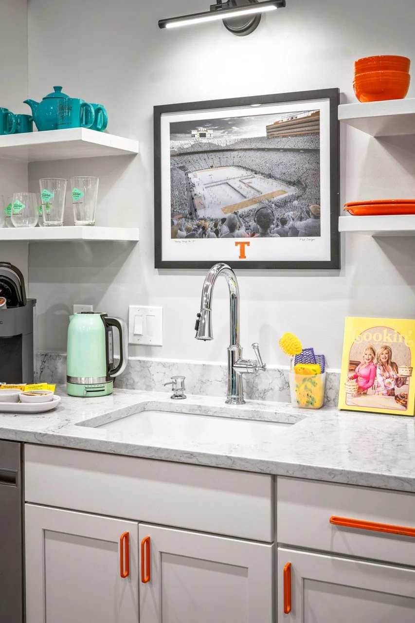 Kitchen sink with a chrome faucet, light green electric kettle, wall-mounted shelves with teal teapots, glasses, and orange dishes, framed photo of a football stadium, cookbook, and kitchen accessories.