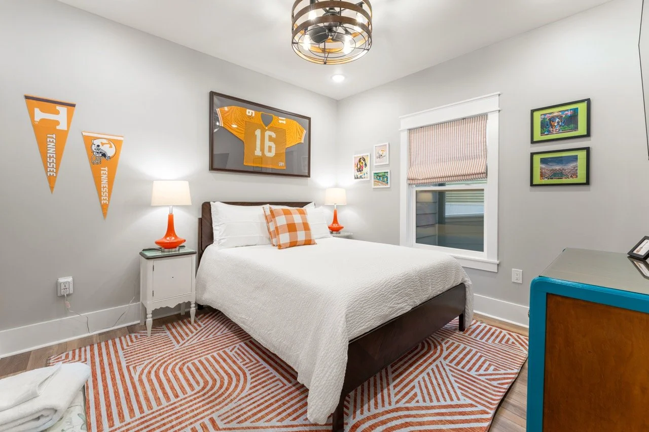A bedroom decorated with Tennessee Volunteers sports memorabilia, including pennants, a framed orange football jersey with the number 16, and framed sports photos, with a bed, white bedding, orange checkered pillow, two orange lamps, and a window wit