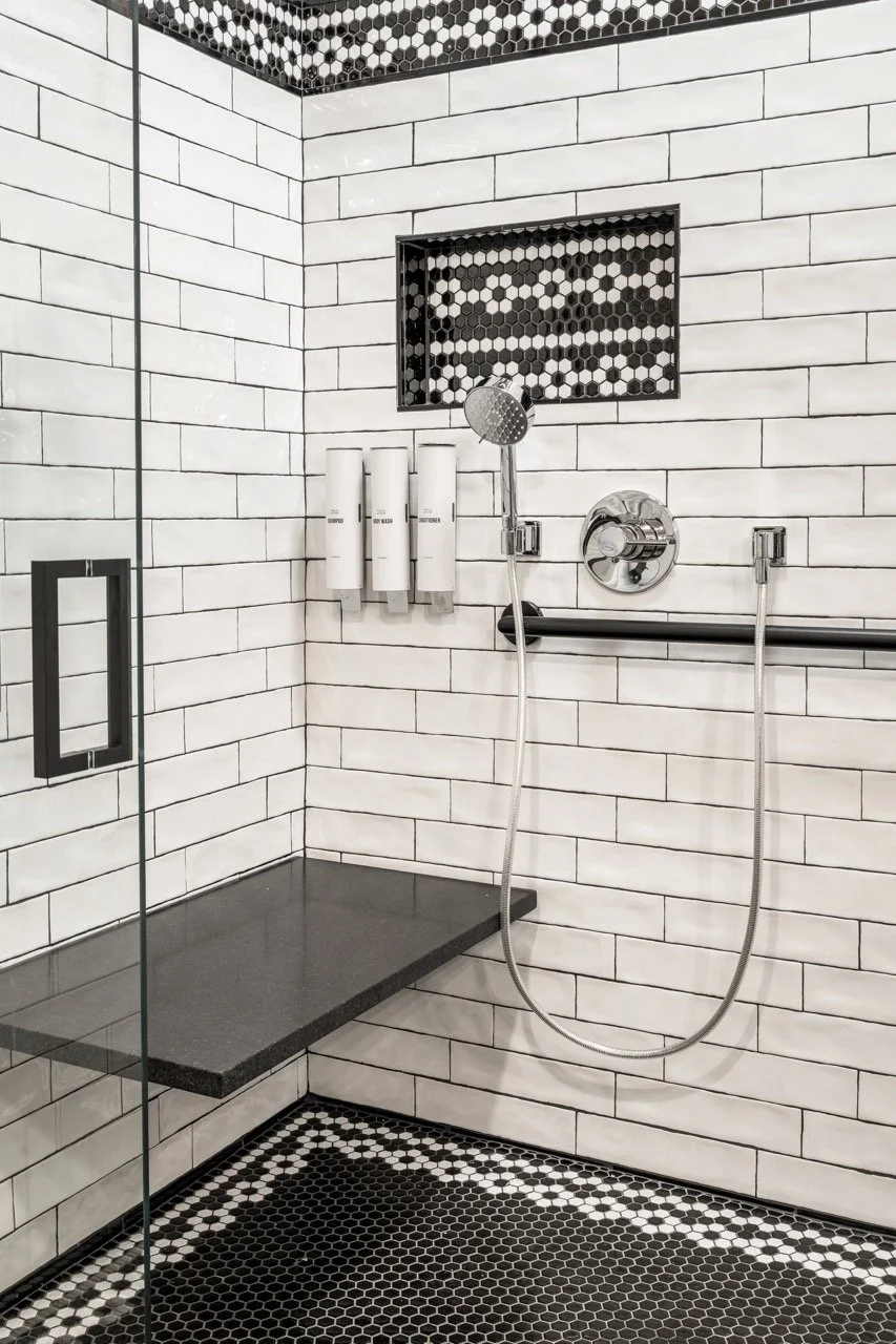A modern shower with white subway tile walls, black hexagonal floor tiles, a built-in shelf with black and white hex tiles, a black bench, a chrome handheld showerhead, and three white dispensers.