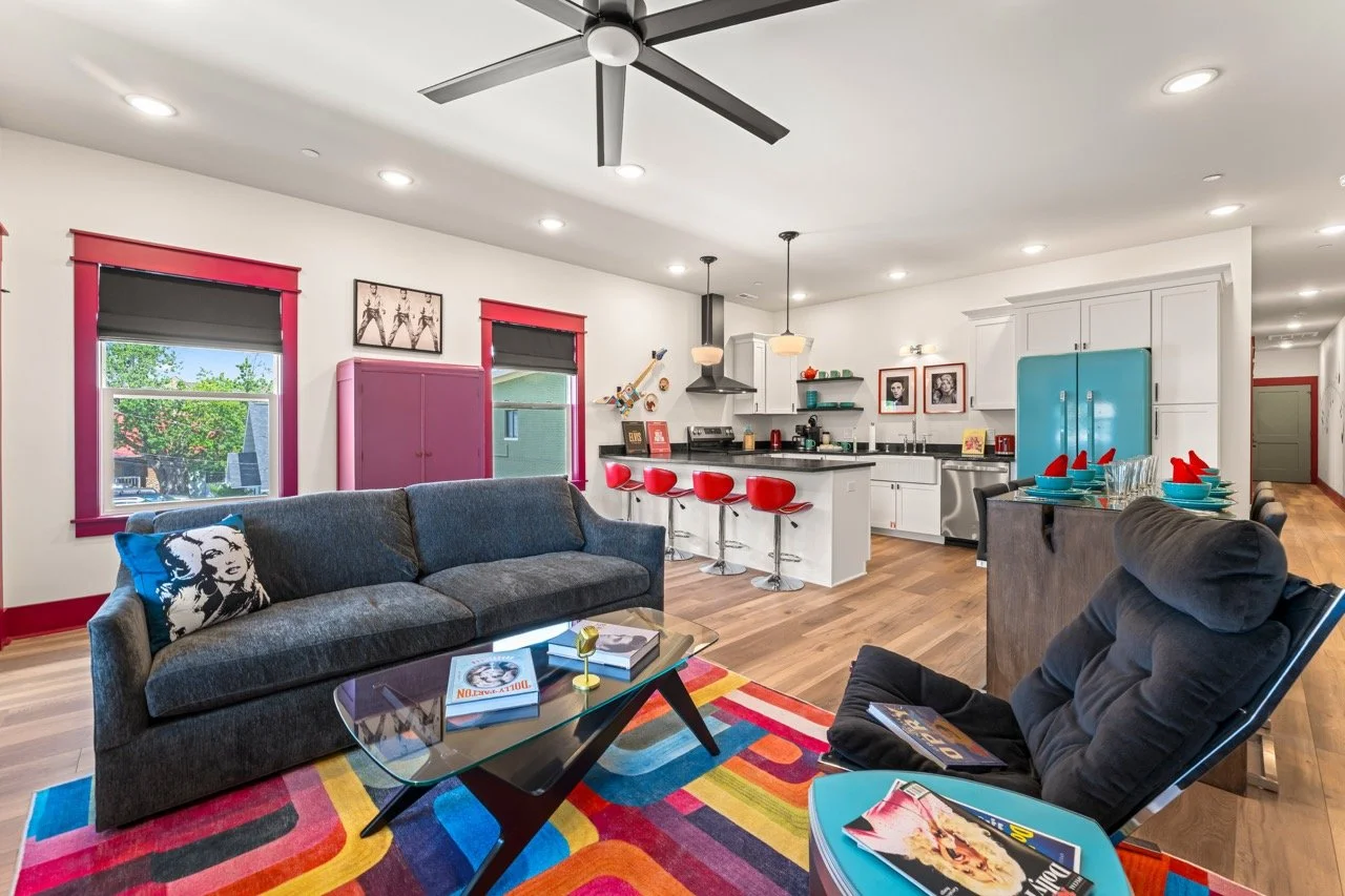 Open-concept living room and kitchen with colorful decor. The living room has a gray sofa, a black armchair, a colorful area rug, and a glass coffee table. The kitchen has white cabinets, a blue refrigerator, red bar stools, and pendant lighting. The