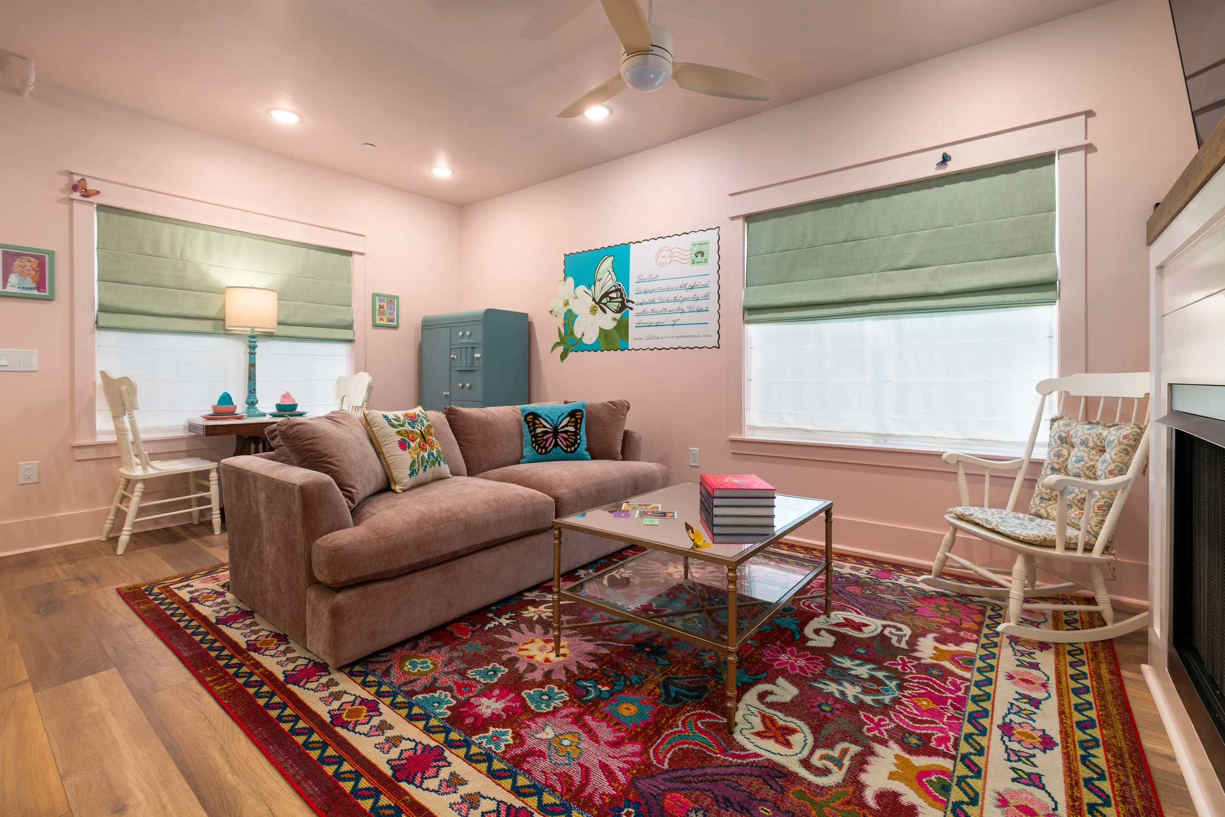 A cozy living room with a colorful patterned area rug, a beige sofa with embroidered pillows, a glass-top coffee table with books, a white rocking chair, a blue cabinet, a floor lamp, and windows with green blinds.