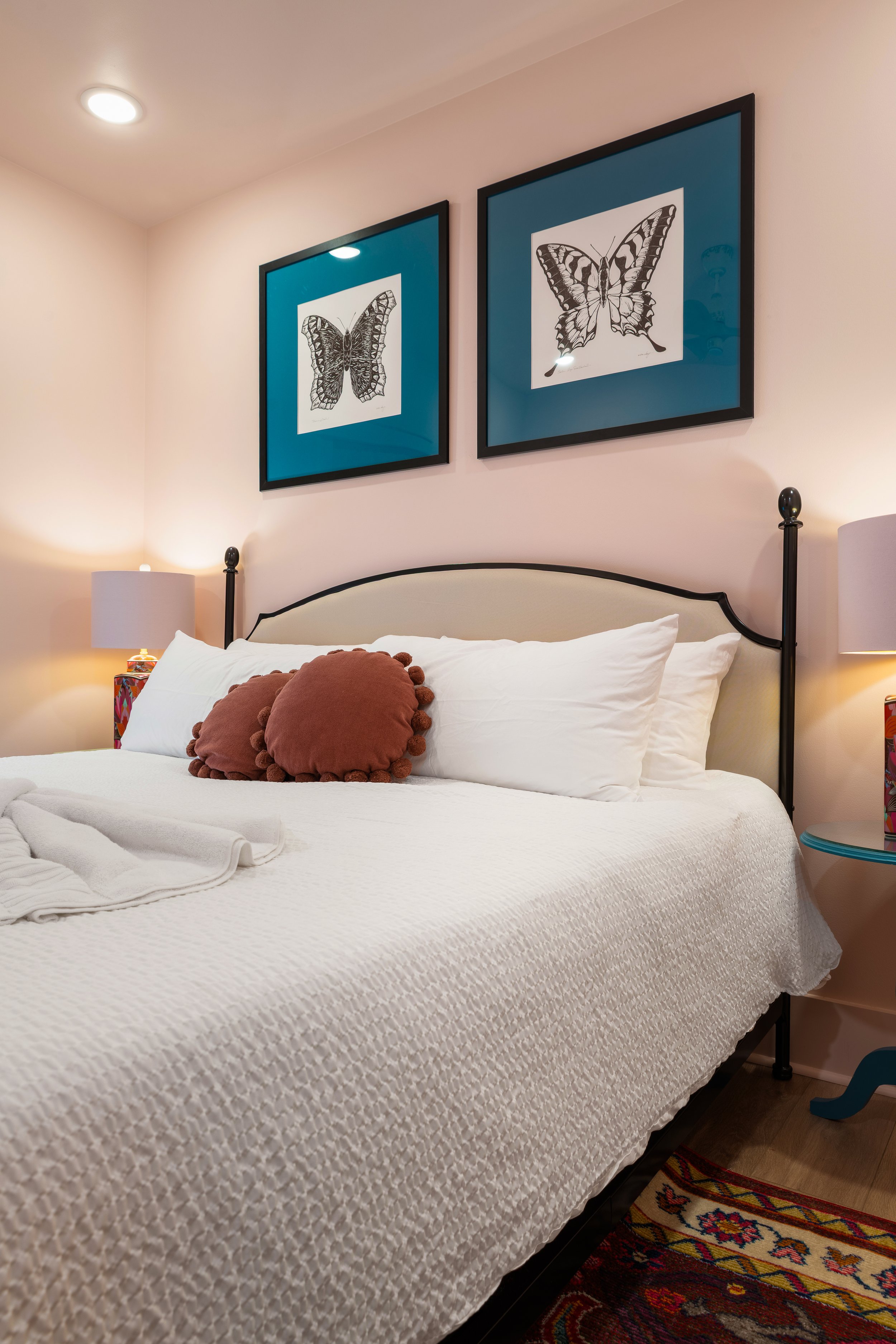 A neatly made bed with a white textured bedspread, decorative brown pillows, and a white towel. Two framed butterfly illustrations are hanging on the wall above the bed, with wall-mounted lamps on each side of the bed, and a colorful patterned rug pa