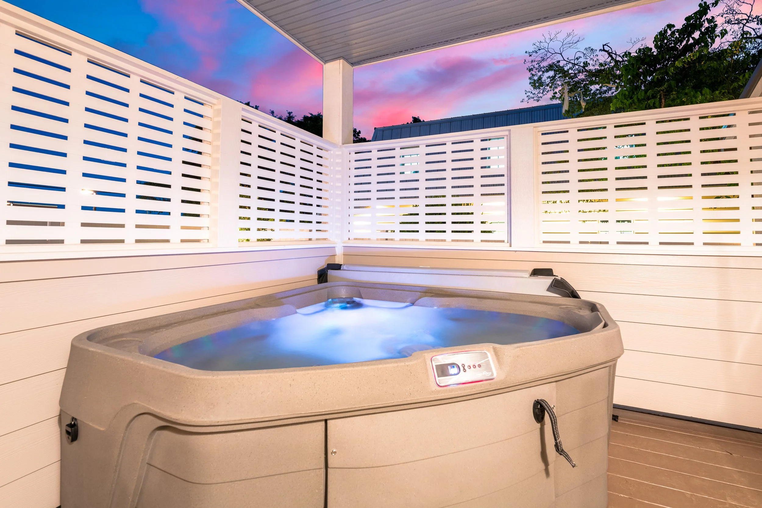 A hot tub is on a porch with white railing and a colorful sky with pink and blue clouds in the background.