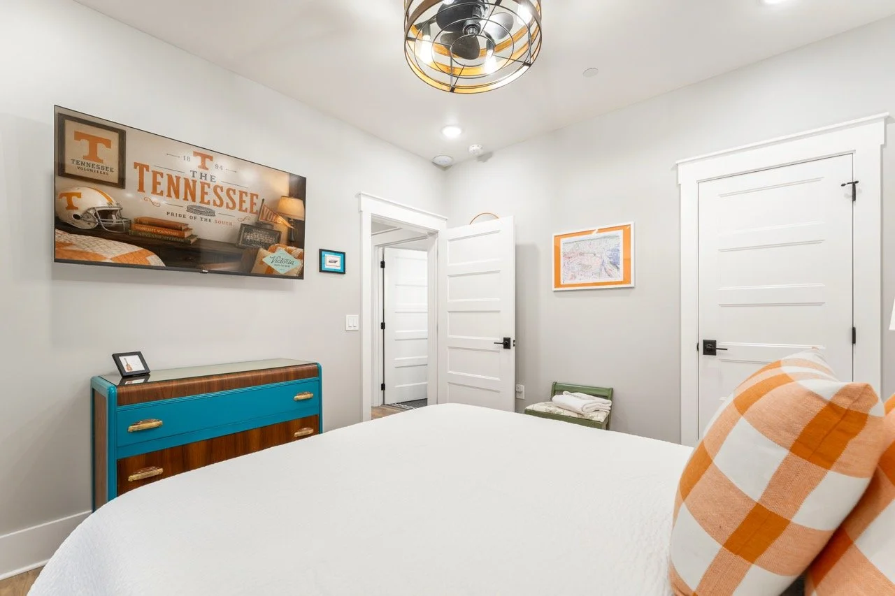 A bedroom with a large bed, white bedding, and orange and white checkered pillows. A blue and wooden dresser is against the wall, and a framed Tennessee Volunteers poster hangs above it. The room has white walls and a modern ceiling light fixture. Th