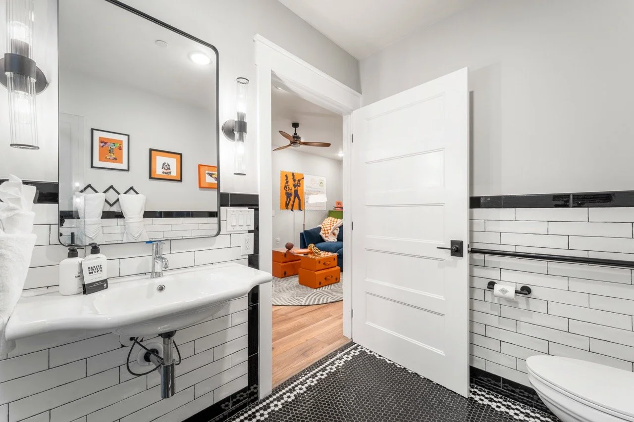 A bathroom with white subway tiles, a white sink with black fixtures, a large mirror, and modern lighting. An open door leads to a living room with a blue sofa, orange storage chests, and wall art.