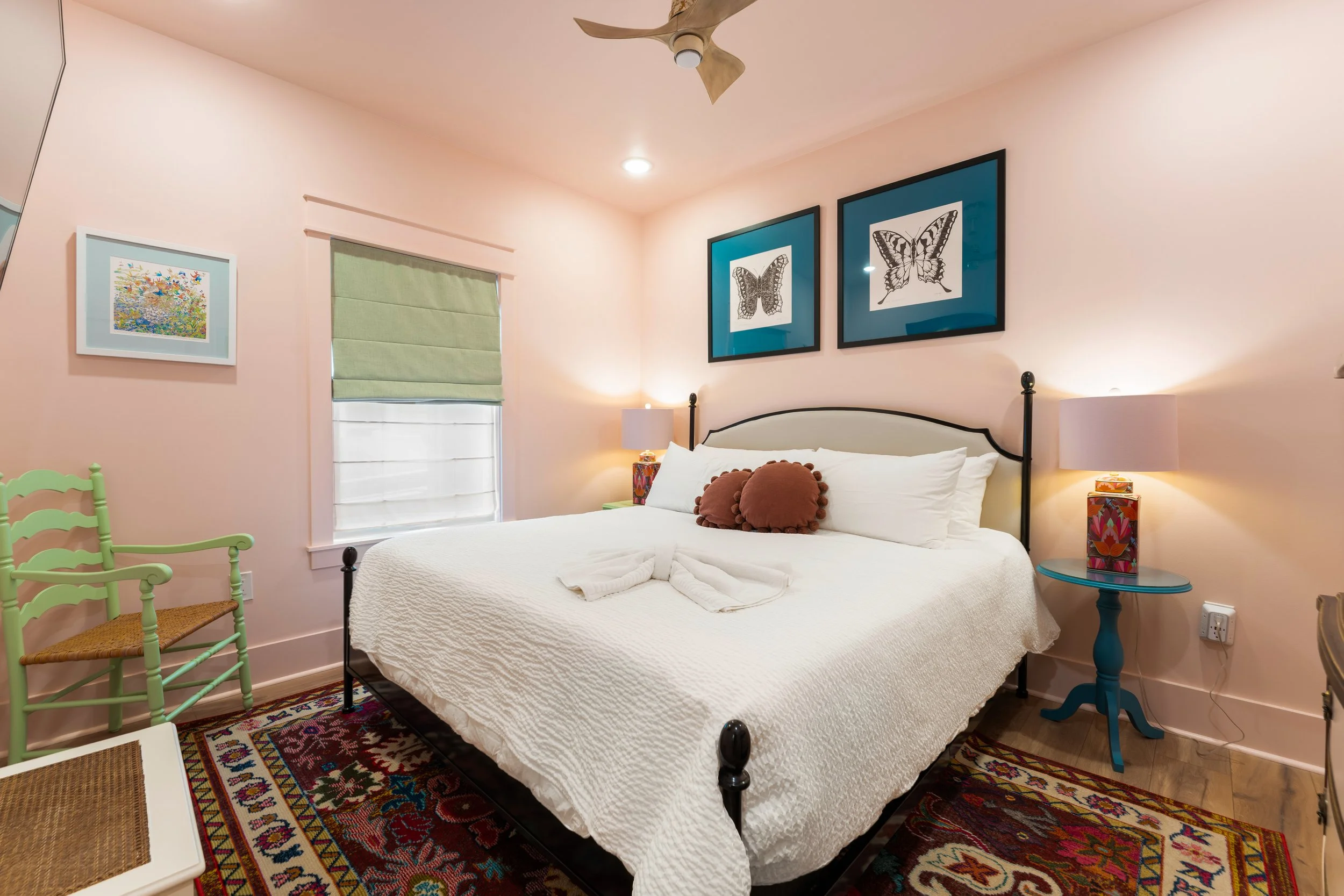A bedroom with a white bed, decorated with two round brown pillows, against a light pink wall. Above the bed, there are two framed butterfly pictures. On the right side, a blue table holds a colorful lamp. To the left, a window with a green Roman sha