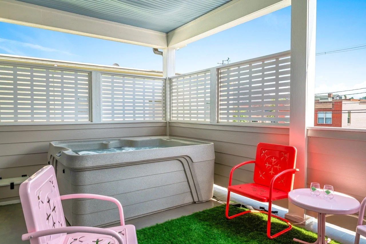Balcony with a hot tub, pink and red chairs, a pink table with two glasses, artificial grass, and a white fence under a blue sky.