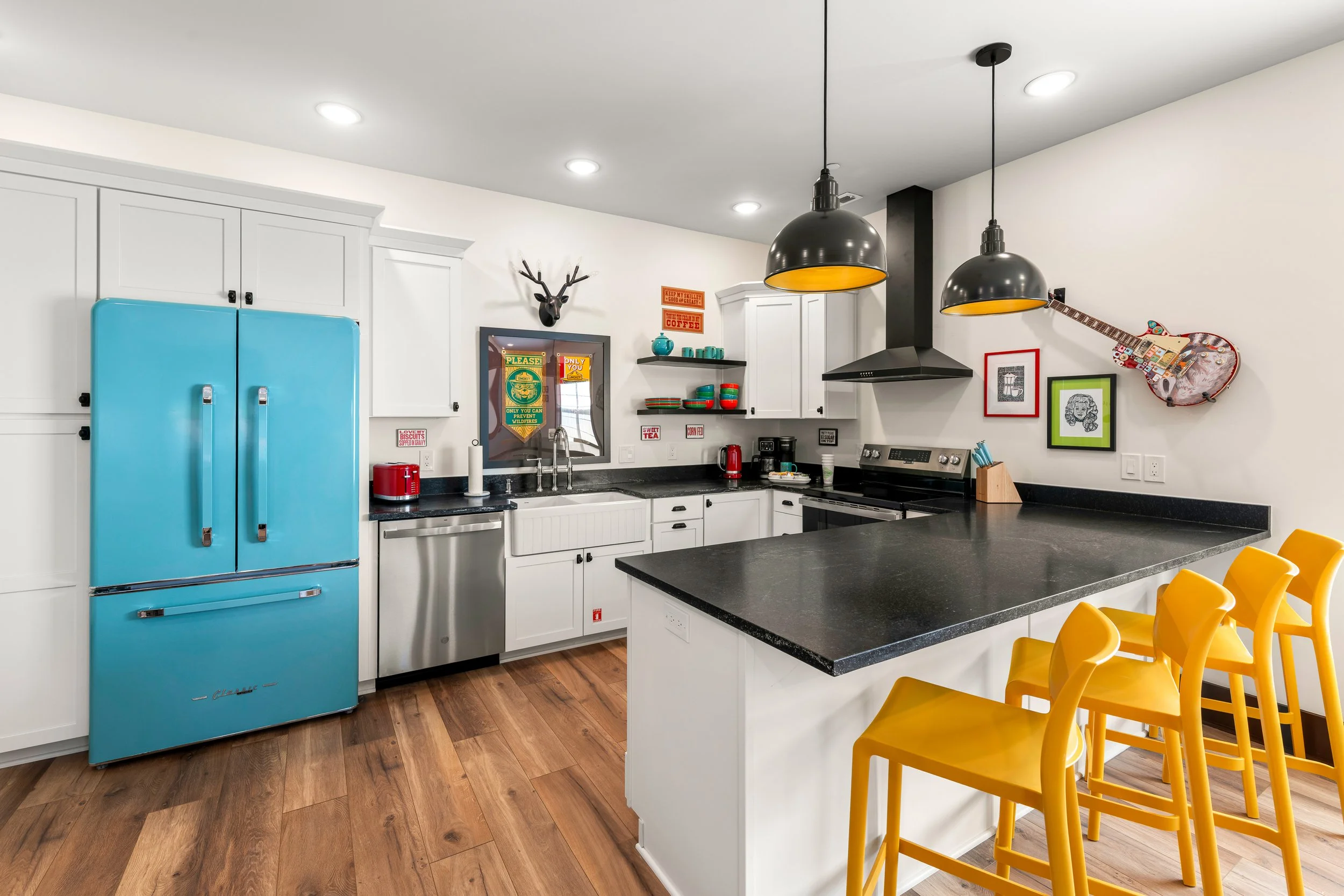 Modern kitchen with white cabinets, black countertops, a bright blue retro-style refrigerator, yellow chairs around an island, open shelving with colorful dishes, black pendant lights, and quirky decor including framed pictures, a deer head wall ornament, and an electric guitar.
