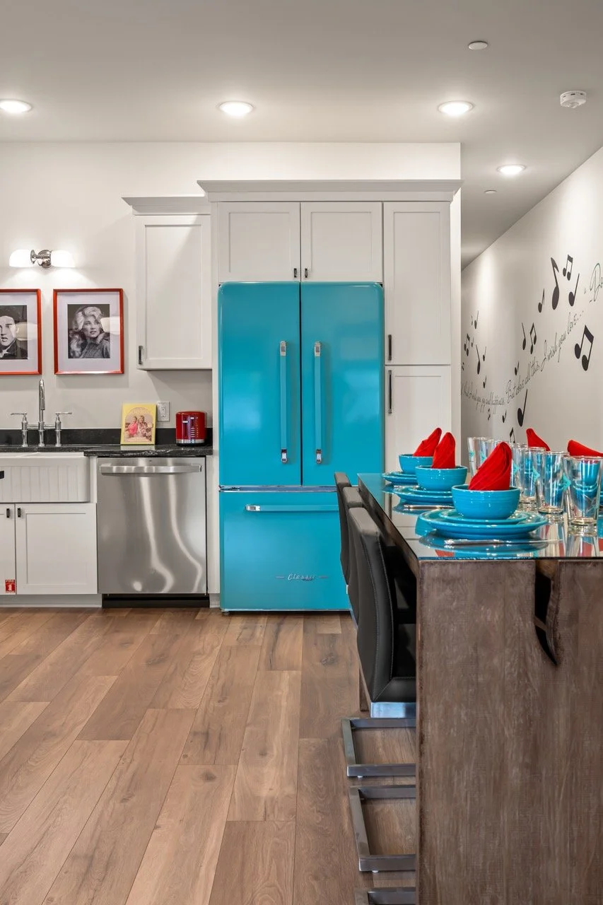 Modern kitchen with white cabinets, a blue retro refrigerator, a stainless steel dishwasher, and a wooden dining table set with red napkins, blue bowls, and glasses. There are framed black-and-white photos and wall art with musical notes.