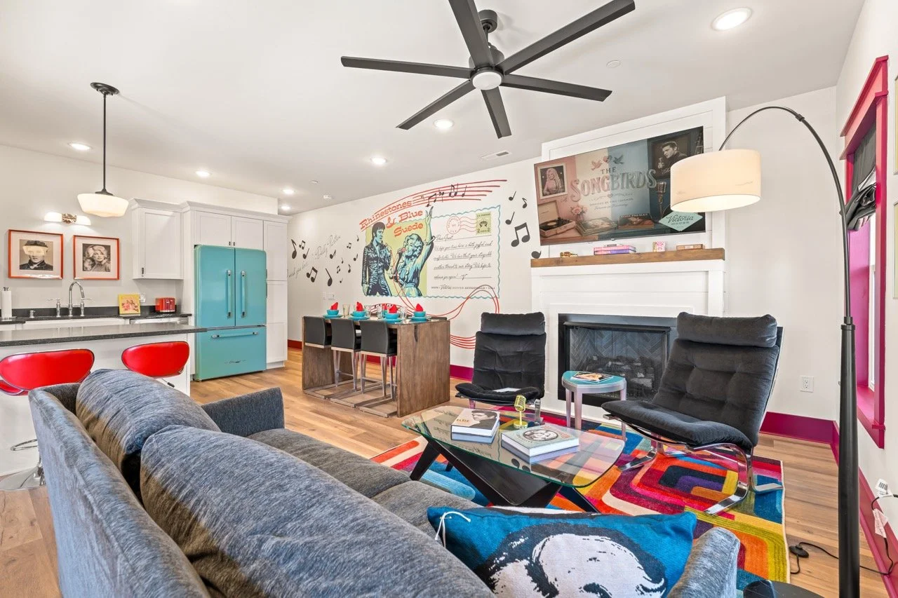 Living room with black chairs, gray sofa, colorful rug, TV above fireplace, and kitchen area with blue refrigerator and red bar stools.