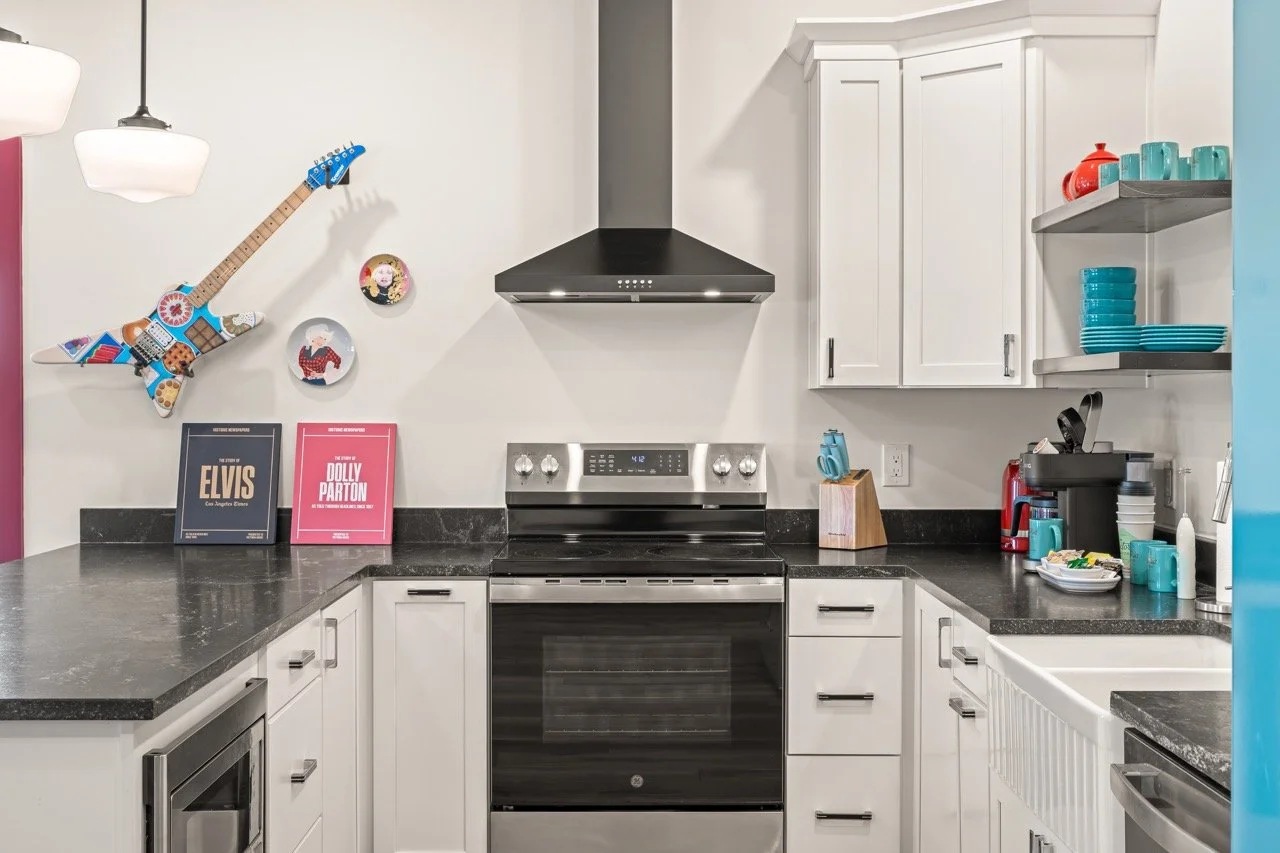 Modern kitchen with black countertops, white cabinets, a black range with digital display, and a black range hood. Decor includes a colorful guitar-shaped wall art and framed posters of Elvis Presley and Dolly Parton. There is a coffee maker, dishes,