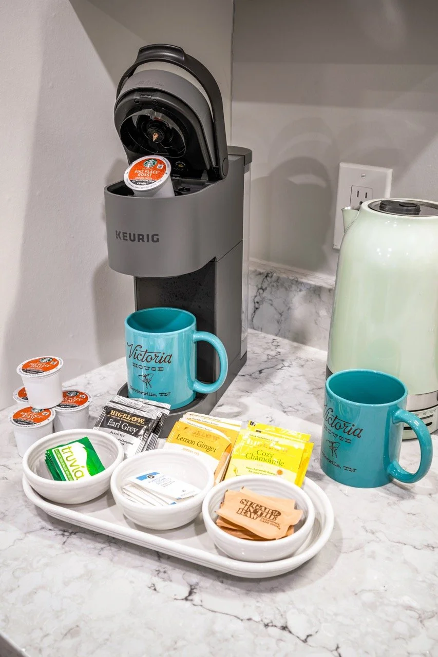 Keurig coffee machine with a brown coffee cup, two blue mugs with 'Victoria' printed on them, various tea and coffee packets, and small cream containers on a marble countertop.