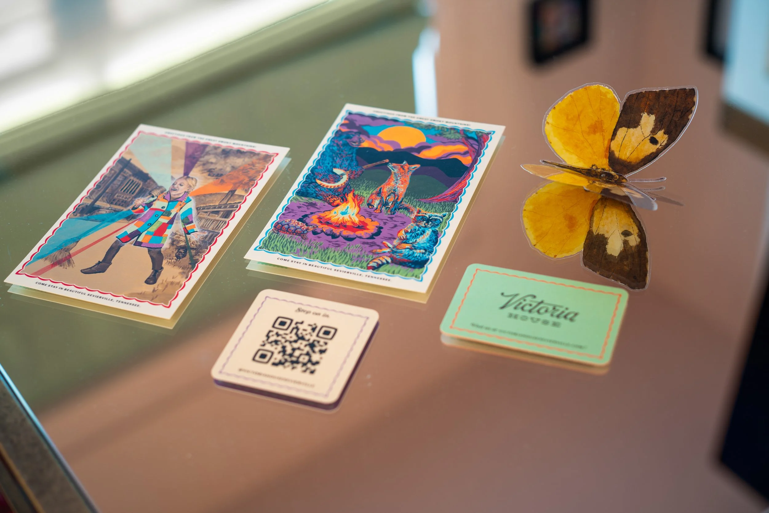 Colorful postcards with illustrations of a woman in a rainbow coat, a mountain landscape with animals, and a butterfly, on a glass surface, along with a QR code and a card with the logo 'Victoria House'.