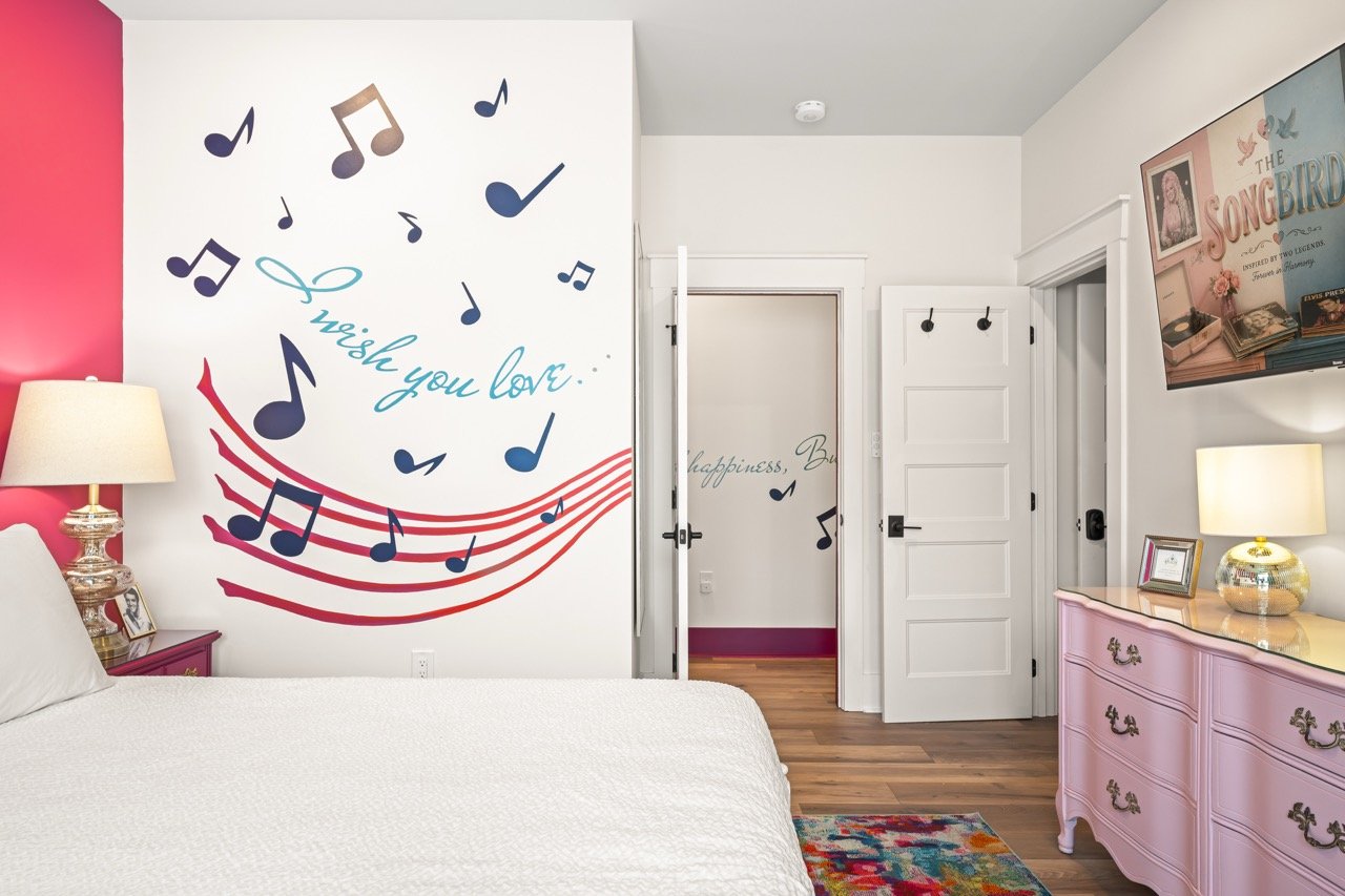 Decorated bedroom with a musical theme, featuring a wall mural with musical notes, a pink dresser, a lamp, a framed photo, and a television.