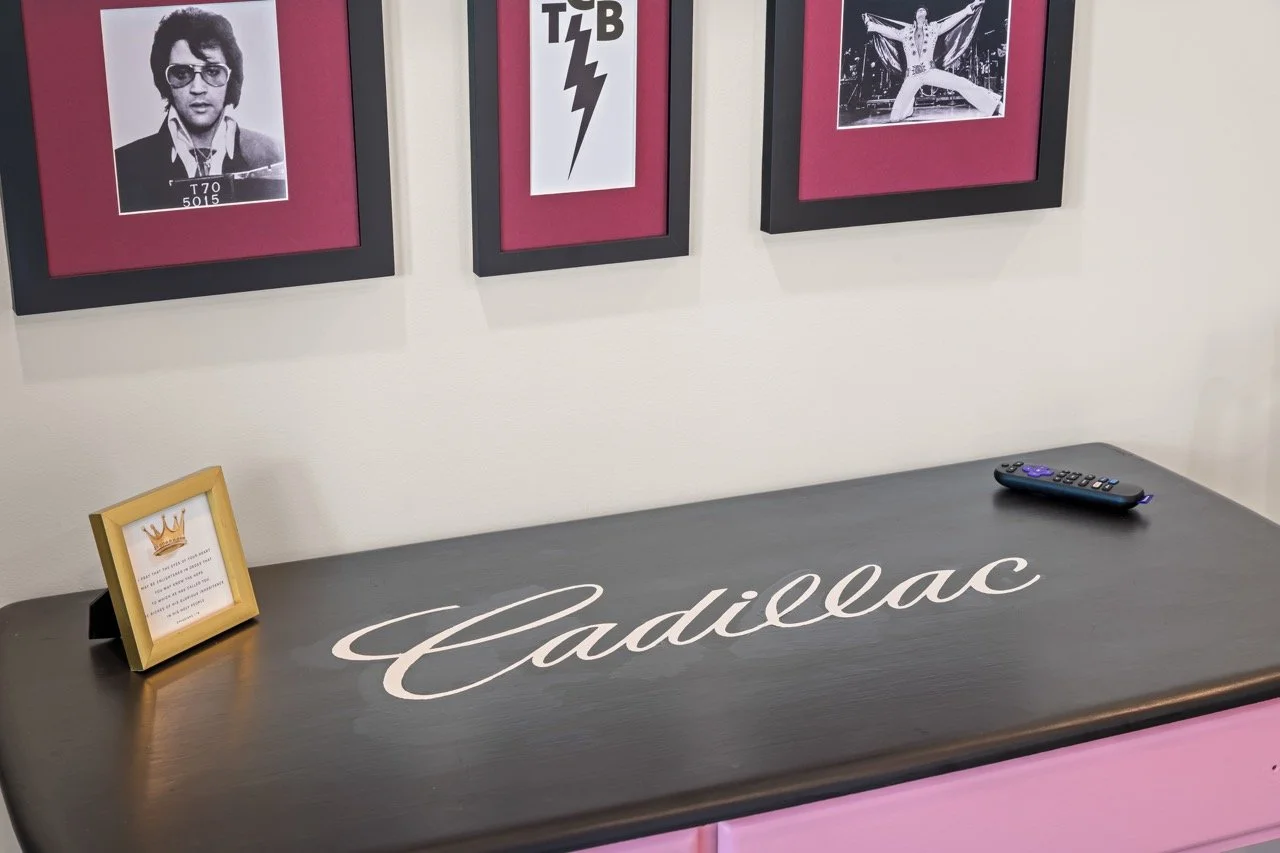 A black desk with the yellow Cadillac logo in cursive on it. On the desk, there is a small framed quote or note and a remote control. Behind the desk, three framed black and white pictures are hanging on the wall, depicting a man with glasses, a ligh