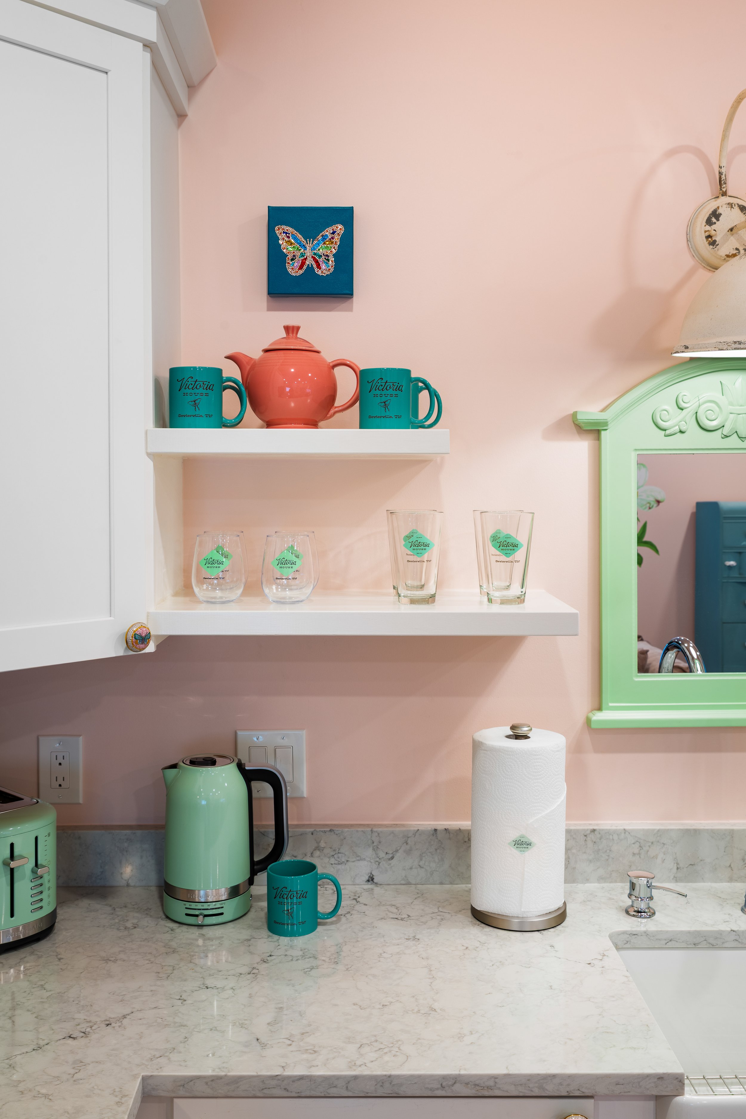 A kitchen counter with a green electric kettle, matching teal mug, paper towel roll, sink, and faucet. Above the counter, there are two teal mugs, a pink teapot, and a blue canvas with a butterfly design. There are also three empty glasses on a white