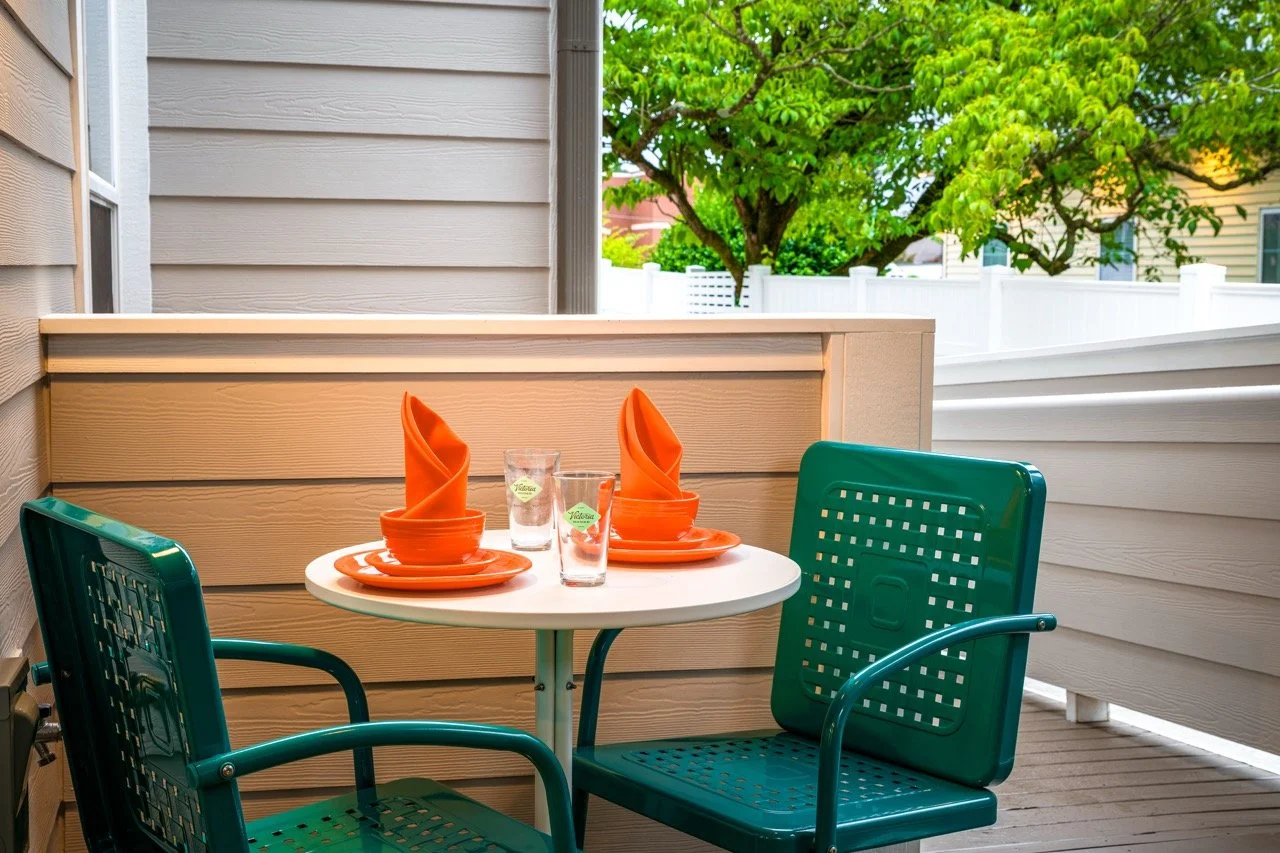 A small outdoor patio with two teal chairs and a white round table set with orange plates, orange napkins, and glasses, with a tree and a white fence in the background.