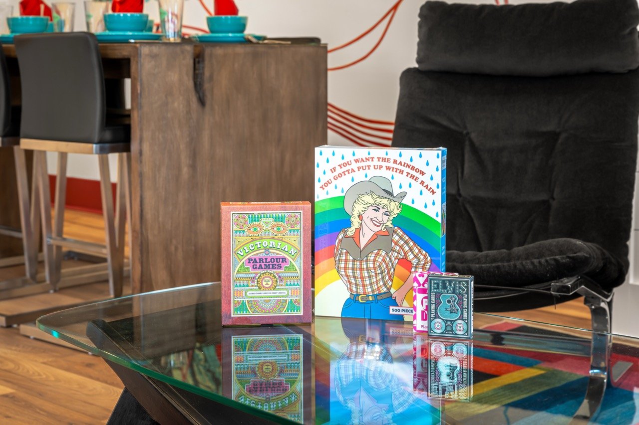 Board game boxes on a glass table, including Victorian Parlour Games, a rain-themed card game with a rainbow and woman in cowboy hat illustration, and Elvis Presley themed playing cards, with a black armchair and a wooden kitchen island with chairs i