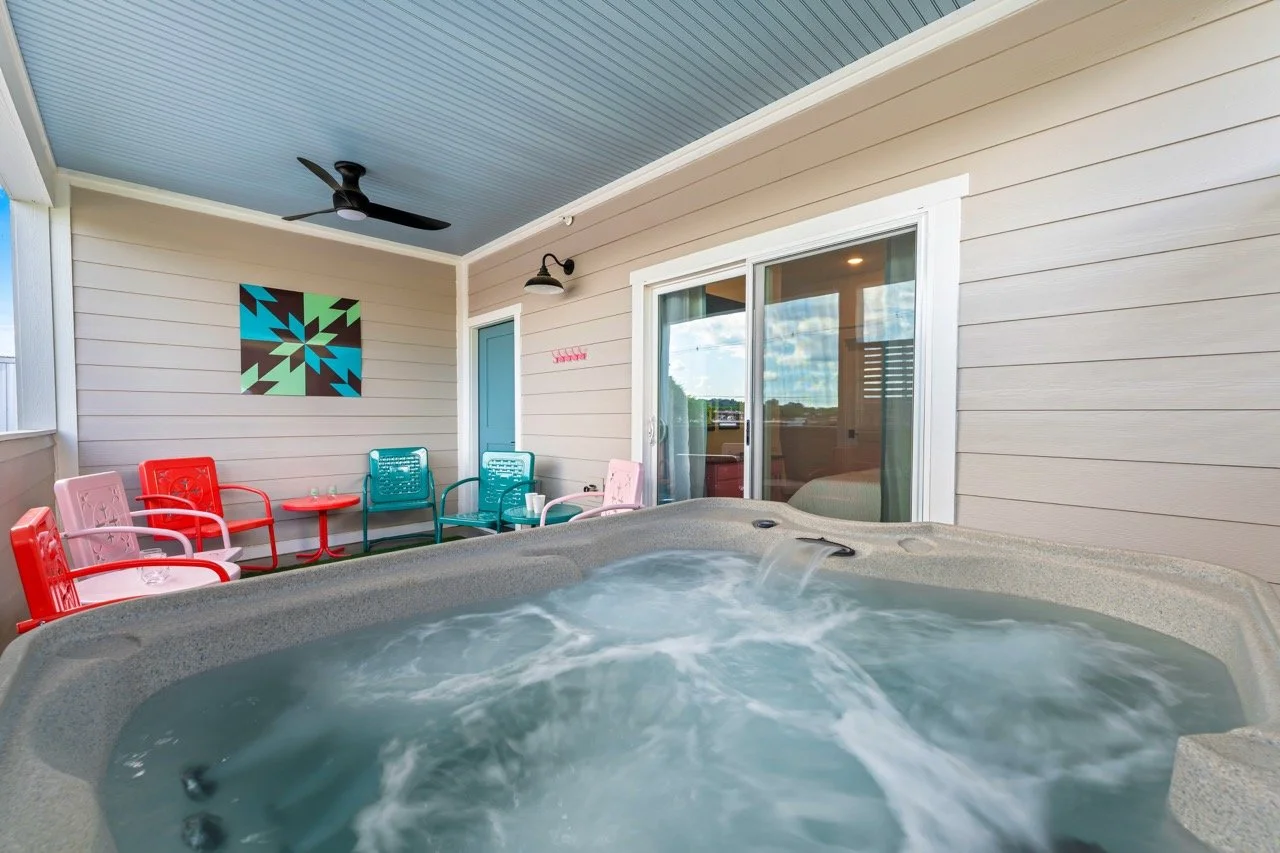 Outdoor porch with hot tub, colorful chairs, small tables, ceiling fan, and wall art, overlooking a partly cloudy sky.