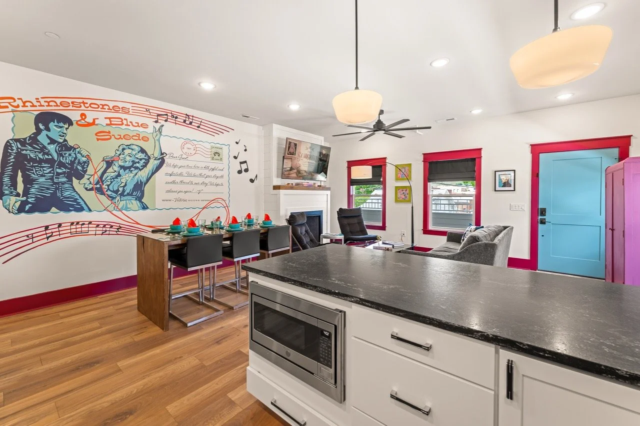 Inside a colorful living room and kitchen area with a wall mural of a singer and a woman holding a microphone. The room has a dining table set with turquoise plates and red napkins, black chairs, a fireplace, a television, and gray and red window fra