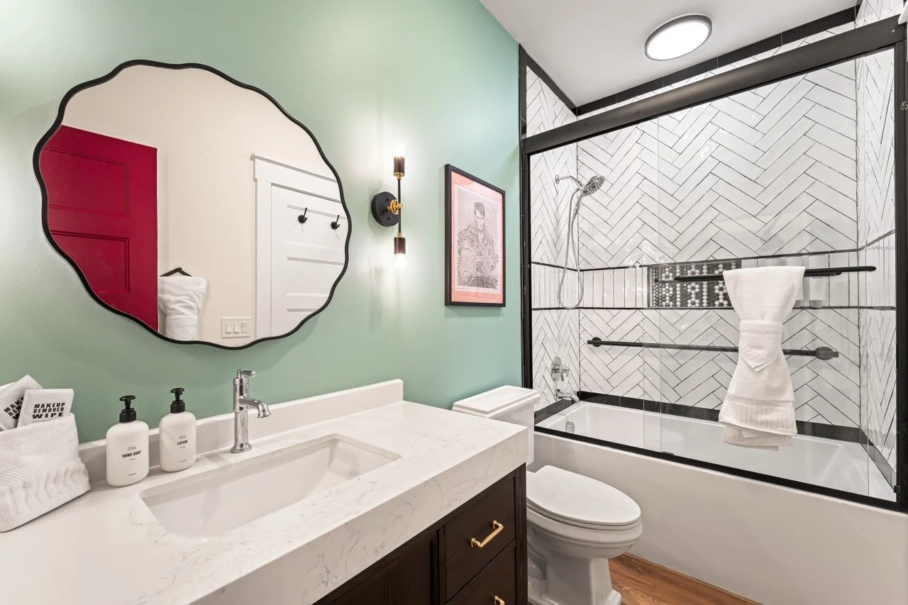 Modern bathroom with green wall, white vanity with marble countertop, oval mirror, black framed shower with white herringbone tile, and a towel hanging in the shower.