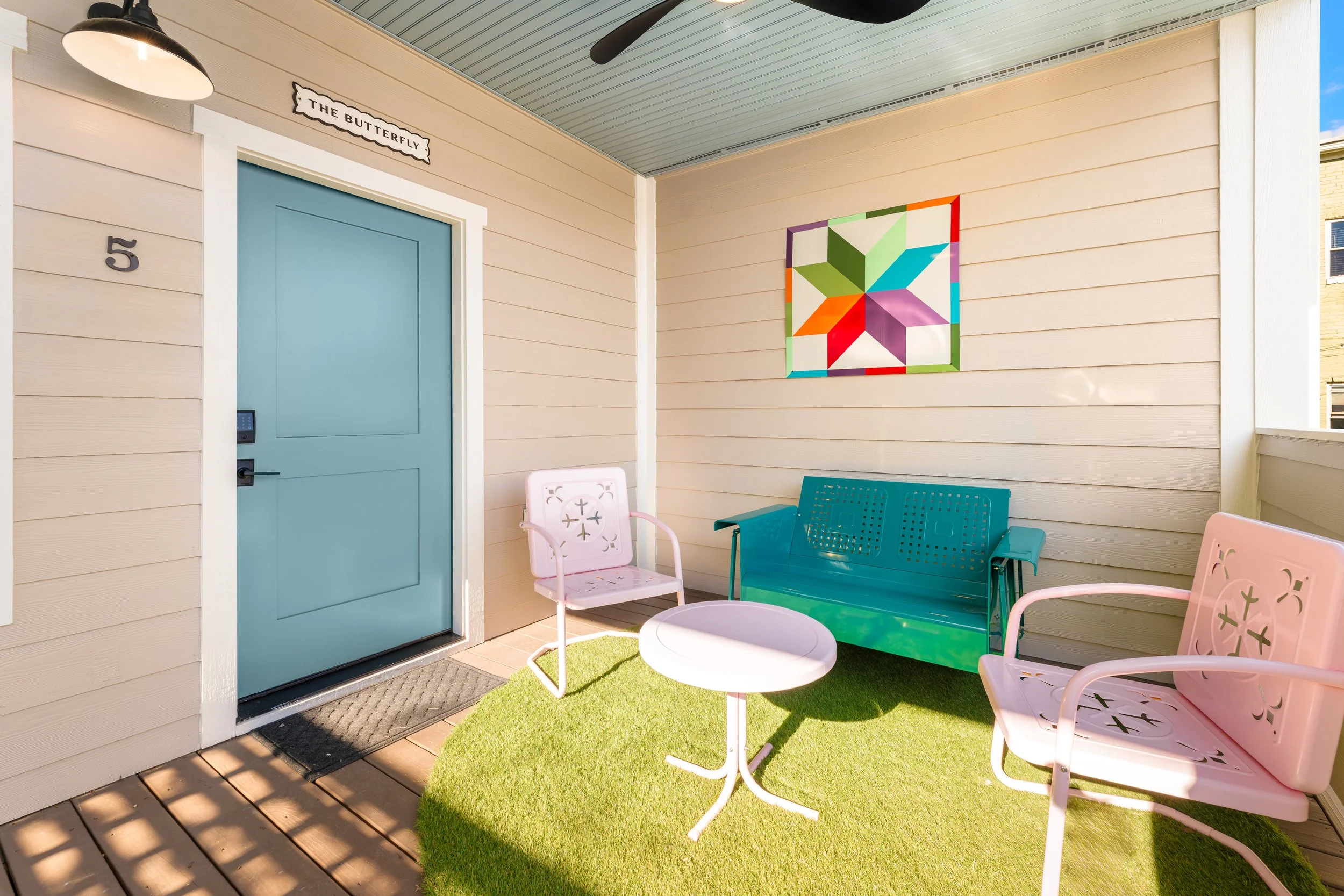 Colorful outdoor porch with pink and blue chairs, small round table, green artificial grass, beige siding, blue door with black handle, wall art, and black outdoor wall light