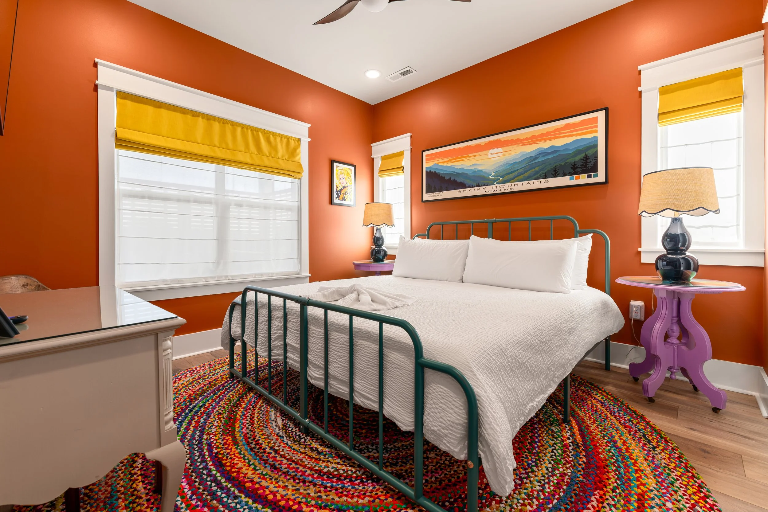 A bedroom with orange walls, white trim, and a multicolored braided rug. There is a bed with white bedding and a green metal frame, flanked by two purple side tables with lamps. Above the bed, there are three framed artworks depicting nature scenes and a portrait of a woman. The room has windows with white blinds and yellow valances, and a ceiling fan.