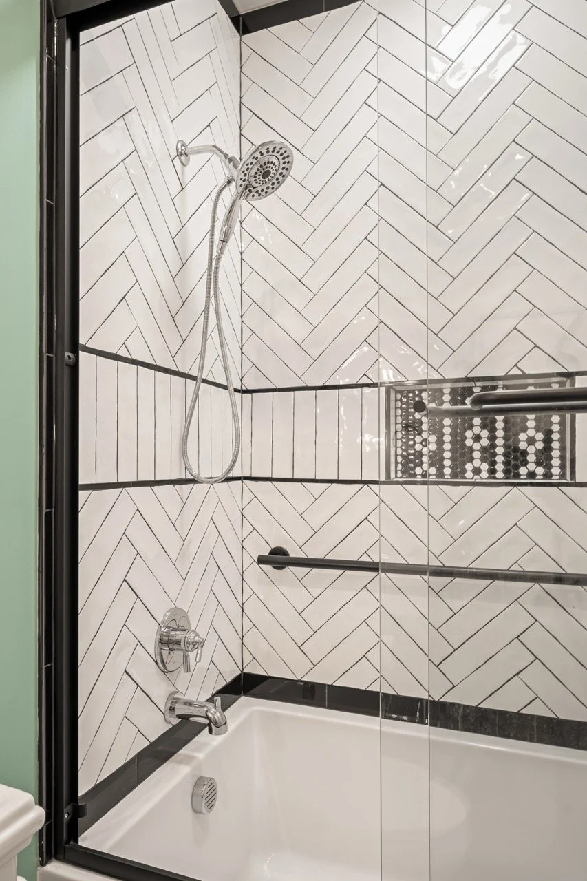 Bathroom shower with white herringbone tile walls, black border tiles, and two black grab bars, with a bathtub below the shower fixtures.