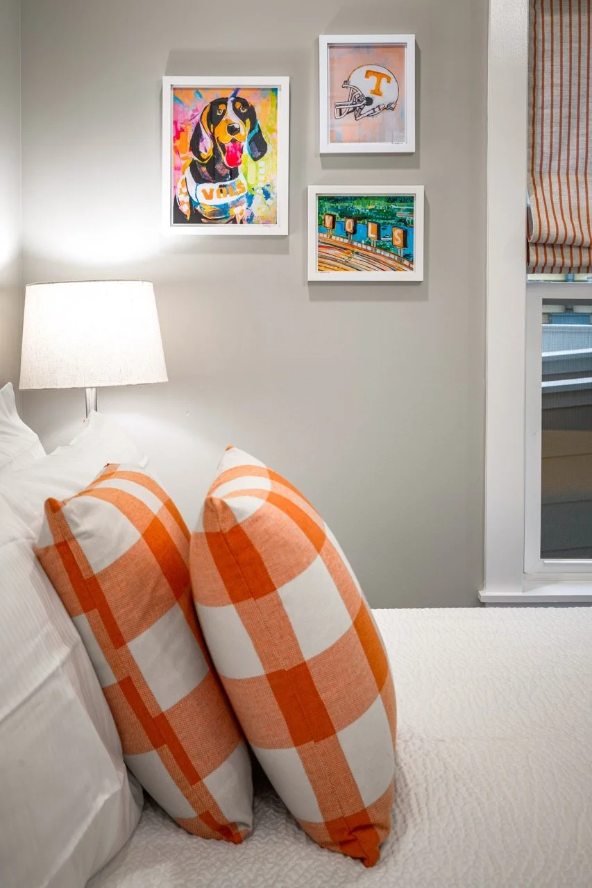 A bedroom corner featuring white and orange plaid pillows on a bed, a white lamp, a light-colored wall with three framed vibrant art pieces, and a window with orange-striped curtains.