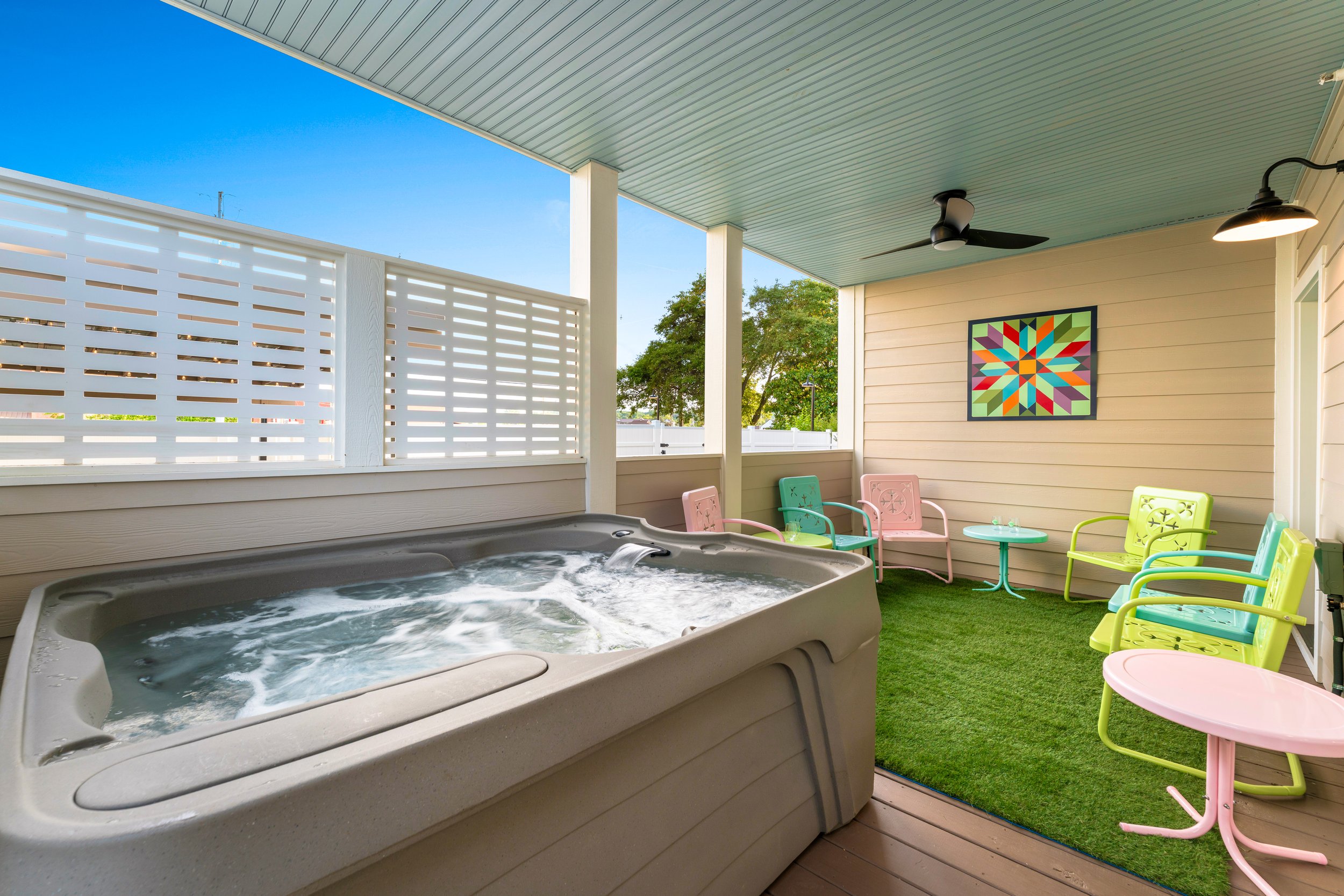 A screened-in porch with a hot tub in the foreground and colorful chairs along the wall. There is a framed geometric art piece on the beige siding, a ceiling fan, and a wall-mounted lamp. The porch has artificial grass flooring and a white lattice privacy fence, with trees visible outside under a clear blue sky.