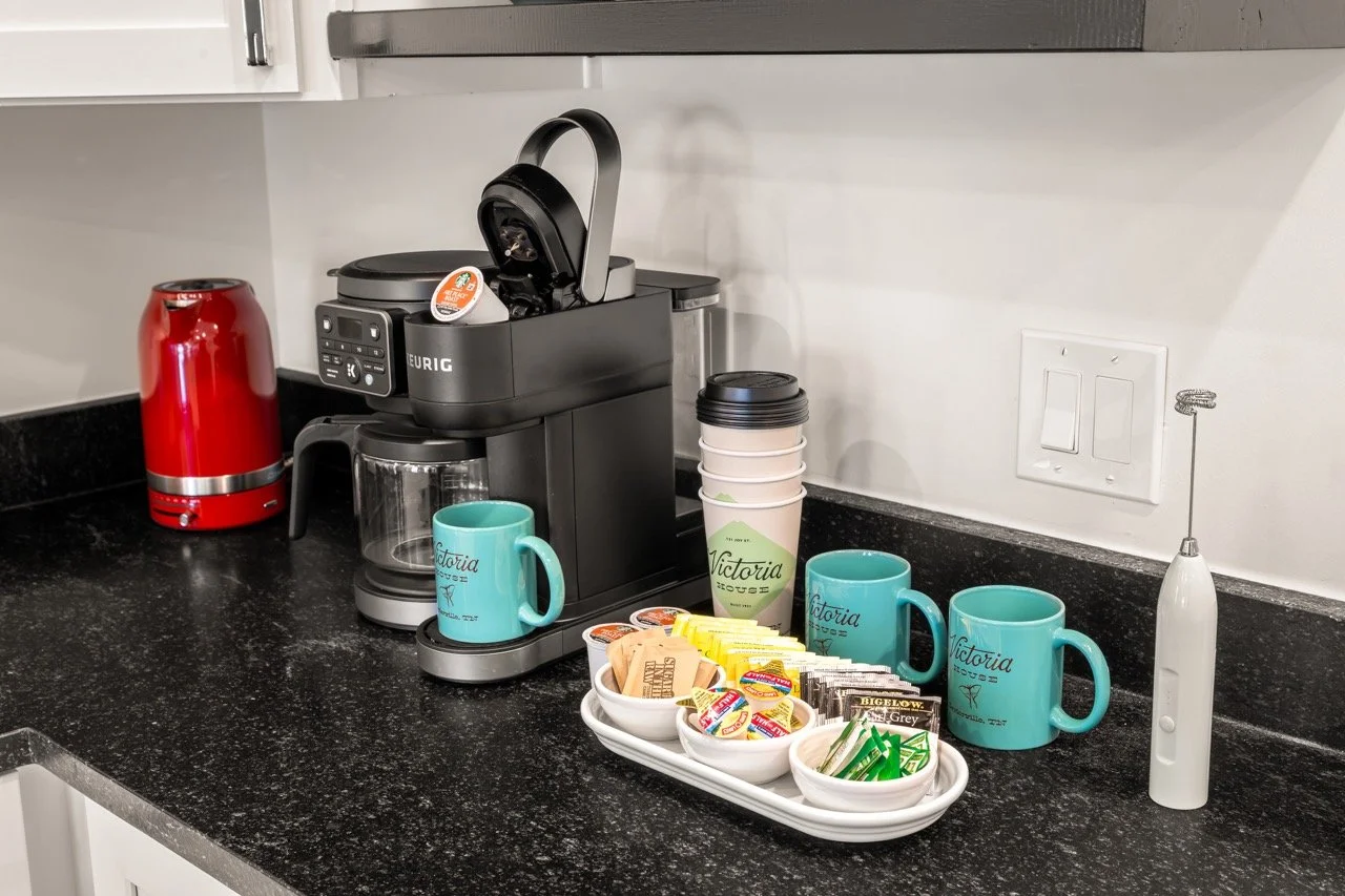 Coffee station on a black countertop with a red thermos, a black coffee machine, three turquoise mugs, a stack of paper cups, and a tray filled with tea bags, creamers, and sweeteners.