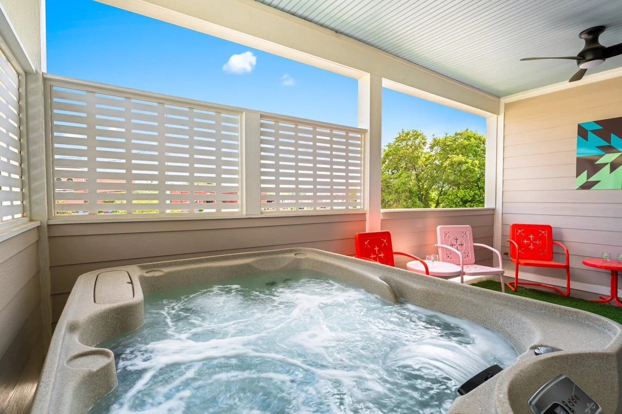 A covered porch with a hot tub, three chairs, and a small table, overlooking trees and a blue sky with a few clouds.