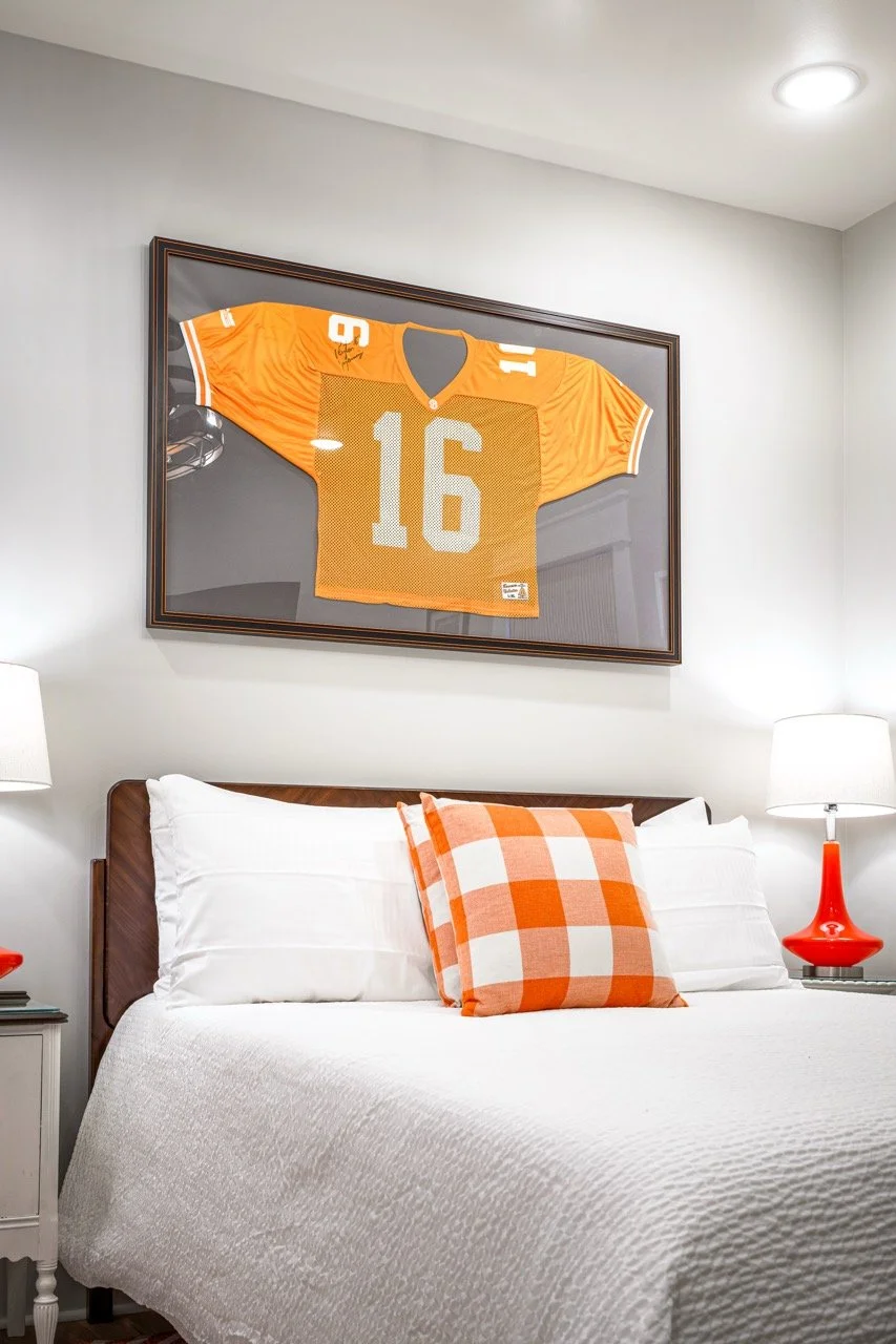 A bedroom with a framed orange football jersey with the number 16 hanging above the bed, which has a white quilt and an orange checkered pillow. There are matching orange and white lamps on bedside tables.
