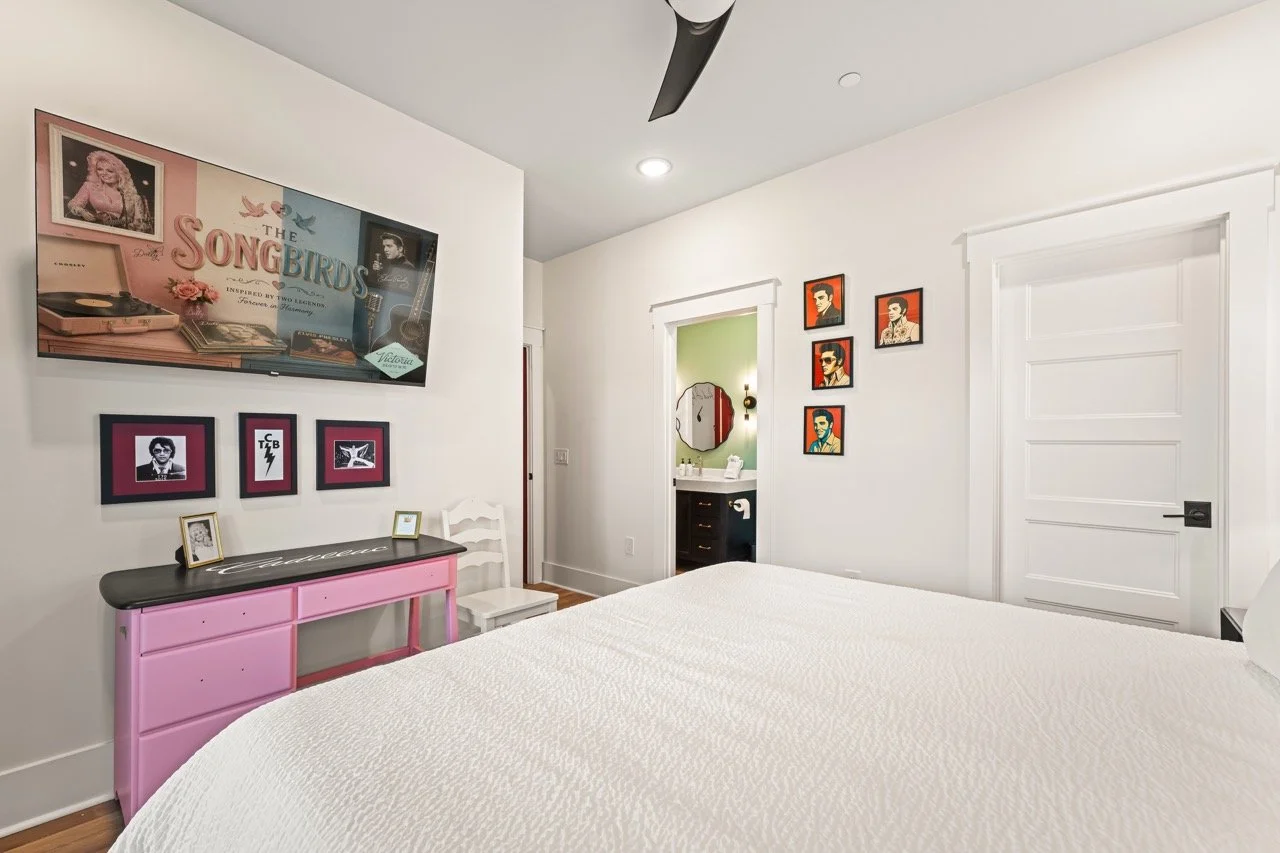 A bedroom with white walls, a white bedspread, and framed photos and artwork on the walls, including a large screen TV displaying a music-themed image, and a pink and black dresser.