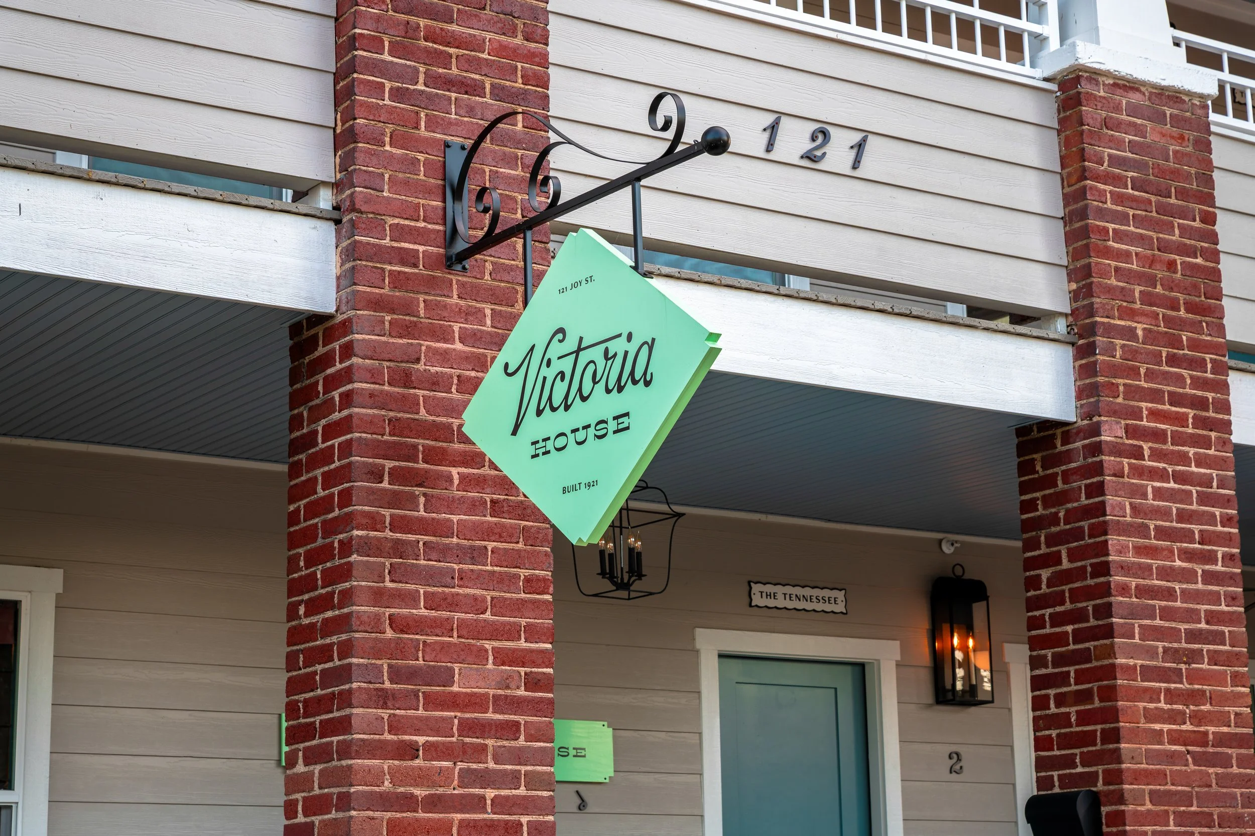 Close-up of a building corner with a brick pillar, beige siding, and a hanging sign reading 'Victoria House,' with addresses 121 Joy St., built in 1921, and decorative black metal elements including a clock-like design.